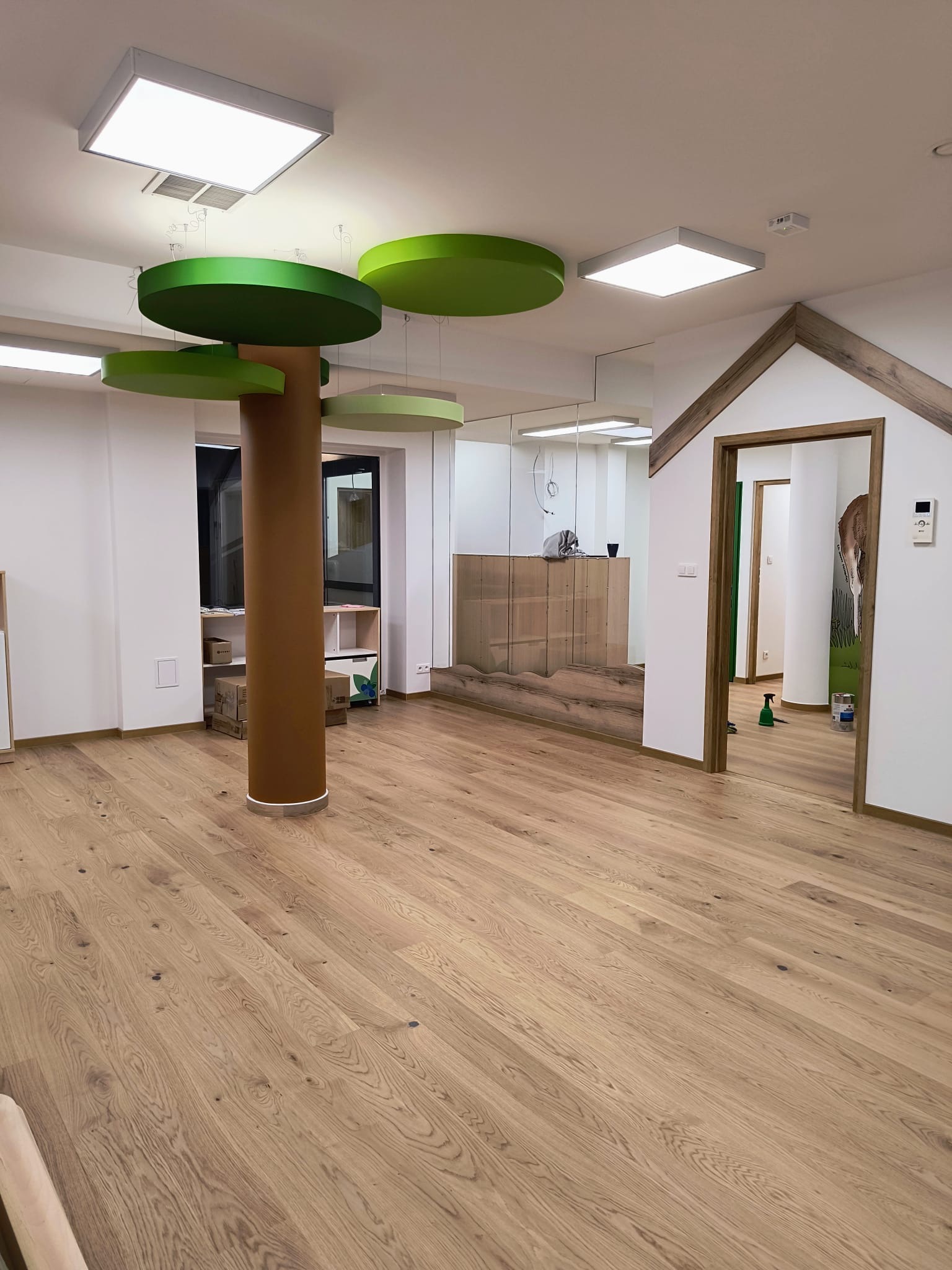Jasna, drewniana podłoga w przestronnym wnętrzu z dekoracyjnym elementem w kształcie drzewa z brązowym pniem i zielonymi okrągłymi liśćmi, widoczne białe ściany i drzwi z drewnianym obramowaniem.
