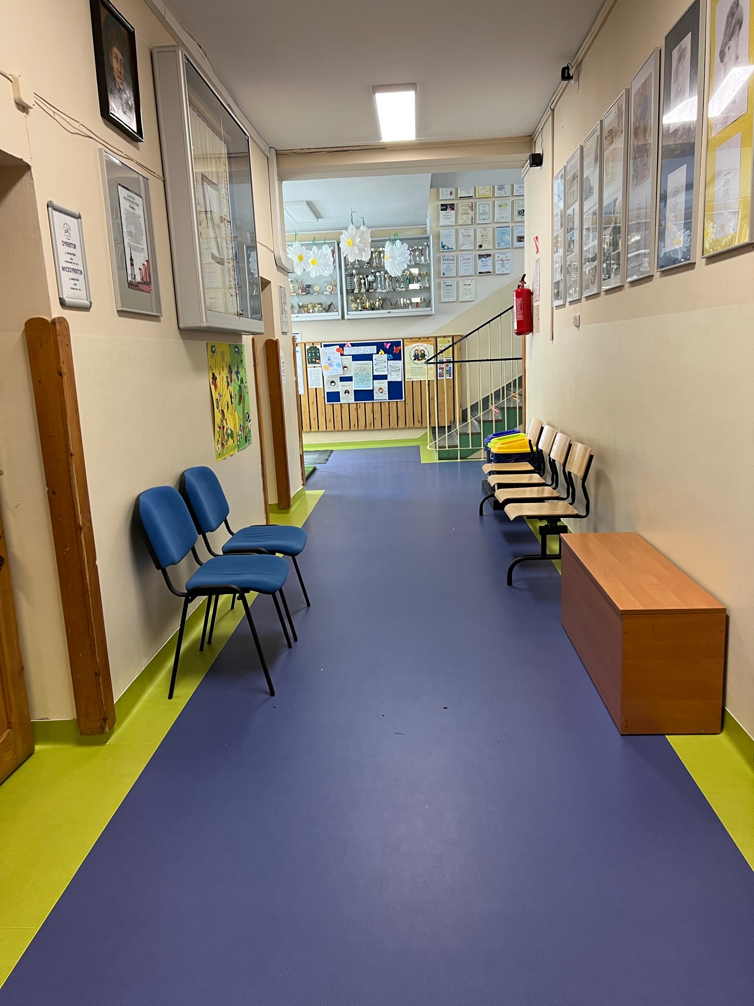 Korytarz szkolny z niebieską wykładziną podłogową, zielonymi listwami przypodłogowymi, rzędem krzeseł i szafką wzdłuż ściany, ozdobiony dyplomami i portretami.