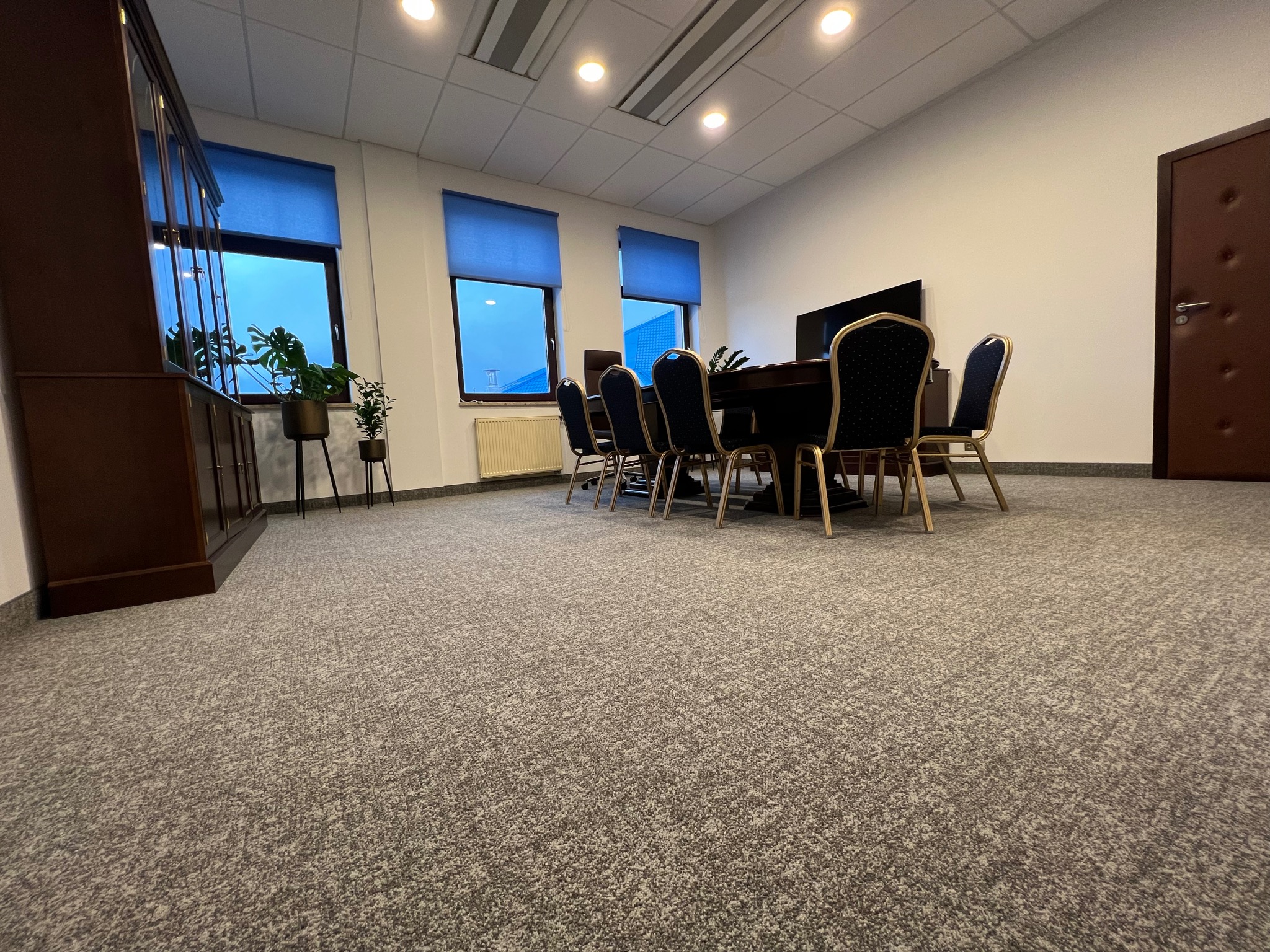 Sala konferencyjna z szarą wykładziną dywanową, ciemnym stołem i krzesłami z czarnymi oparciami, widok z niskiej perspektywy.