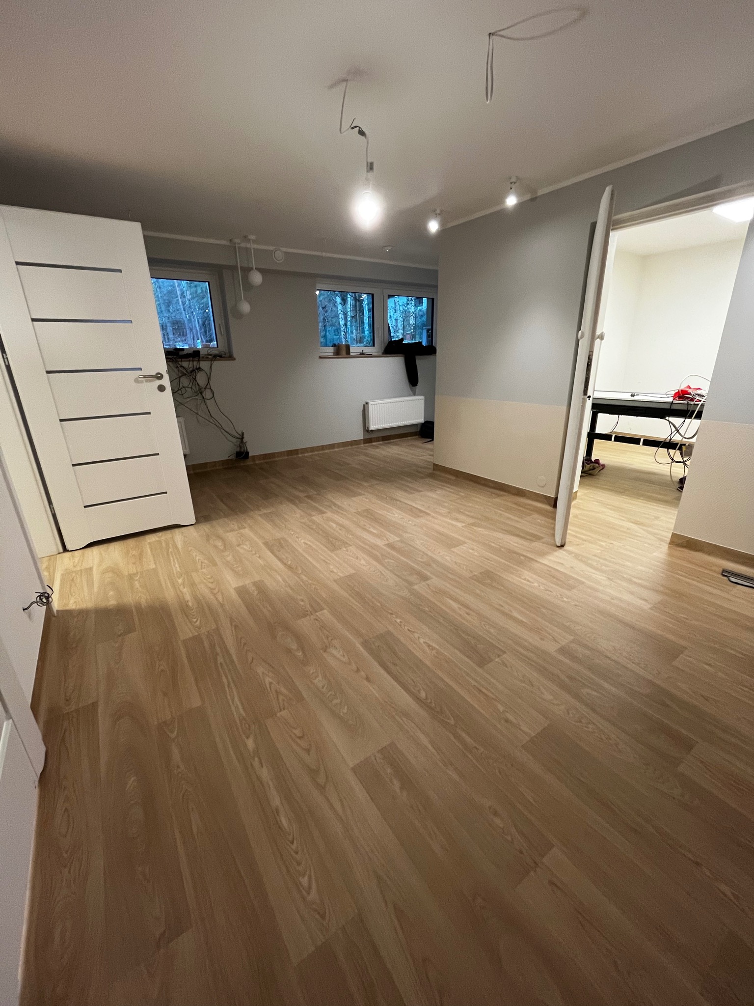 Świeżo ułożona, jasna podłoga w pokoju z otwartymi drzwiami, widoczne okna z widokiem na drzewa i grzejnik pod oknem.