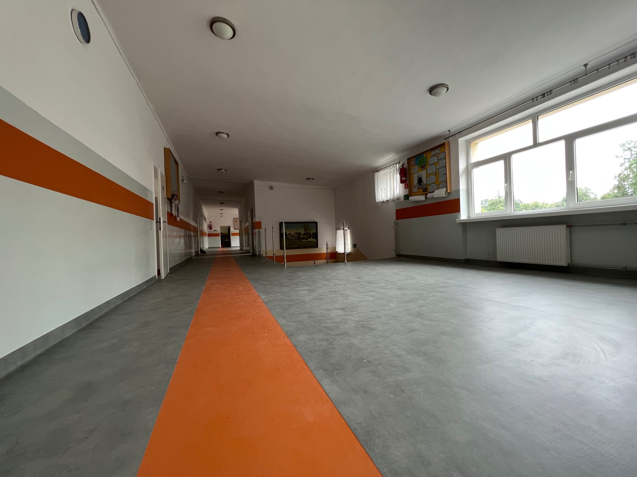 Długi korytarz w budynku publicznym z szarą posadzką i pomarańczowym pasem biegnącym przez środek, białe ściany z szarym i pomarańczowym paskiem, okna po prawej stronie, widok na zewnątrz.