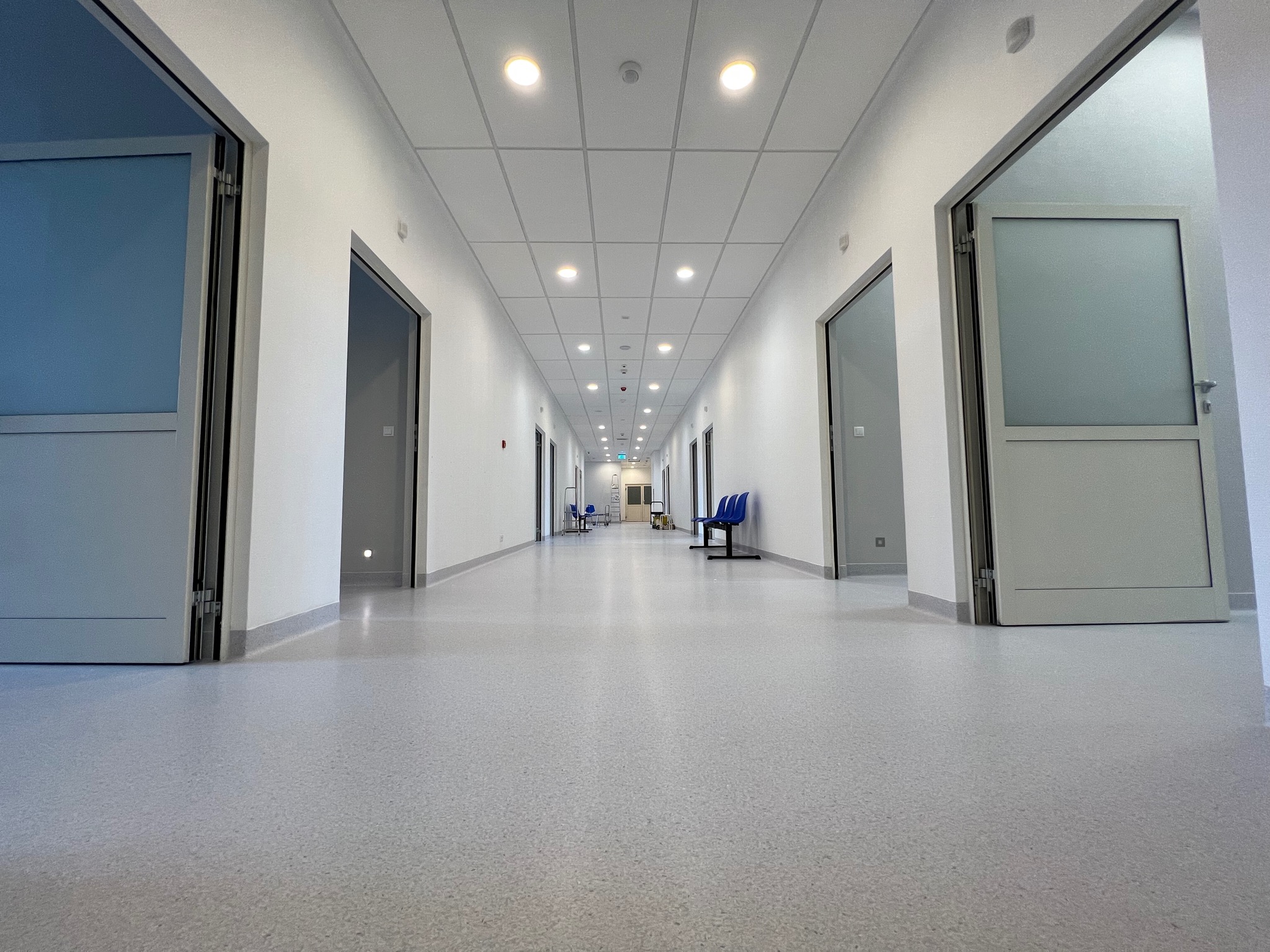 Długi, jasny korytarz w szpitalu lub klinice z szarą podłogą, białymi ścianami i rzędem zamkniętych i otwartych drzwi po obu stronach, oświetlony wbudowanymi lampami sufitowymi.