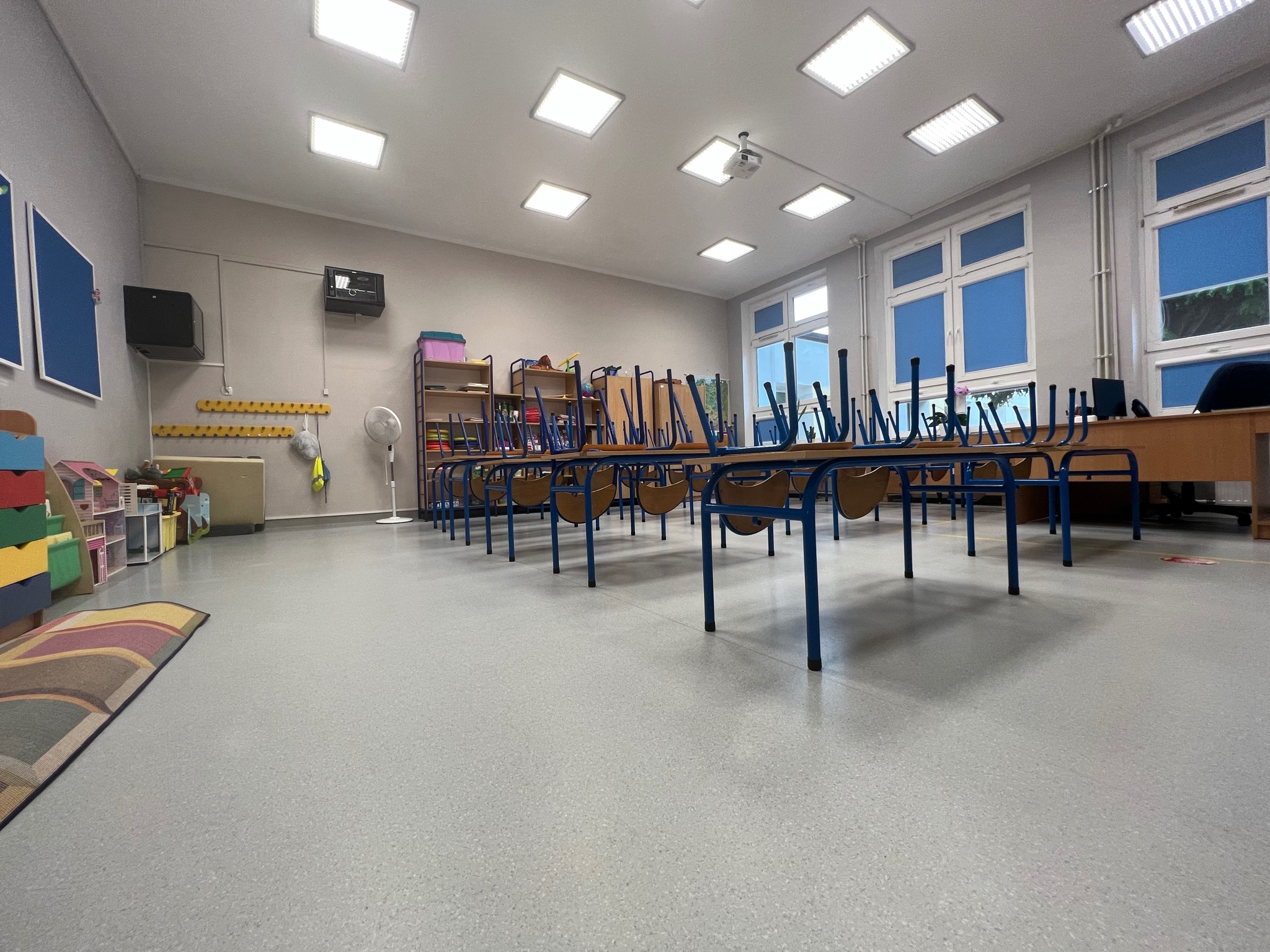 Szara, bezspoinowa posadzka w sali przedszkolnej z krzesłami na stołach po sprzątaniu, widoczne okna i oświetlenie sufitowe.