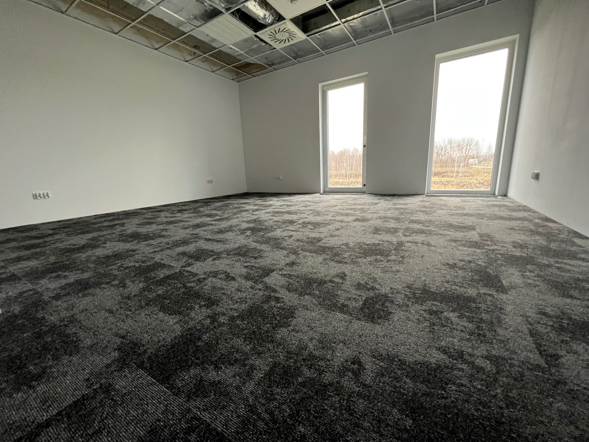 Nowo ułożona, ciemnoszara wykładzina dywanowa w płytkach w pustym biurze w Warszawie, widoczne okna z widokiem na drzewa i sufit podwieszany.