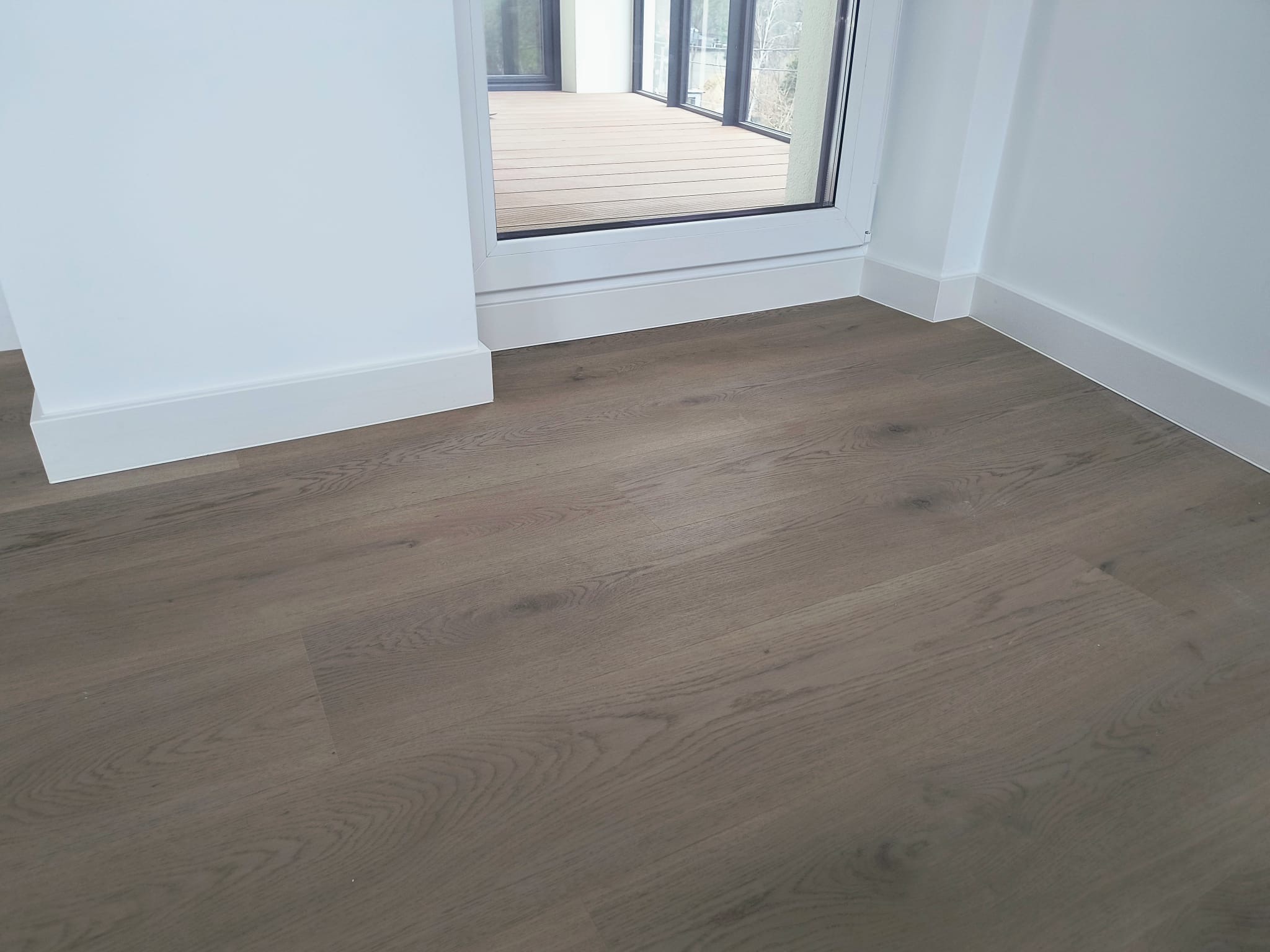 Ułożone panele podłogowe w odcieniach szarości, widoczne białe listwy przypodłogowe i fragment okna balkonowego.