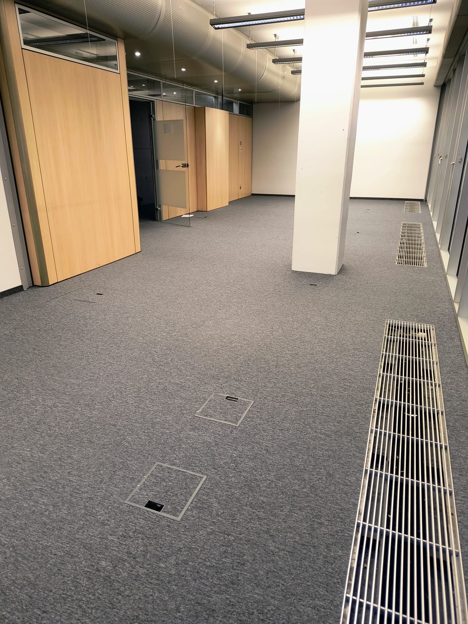 Szara wykładzina dywanowa w biurze z kratkami wentylacyjnymi i podłogowymi gniazdami elektrycznymi, widok na przeszklone biura z drewnianymi drzwiami.