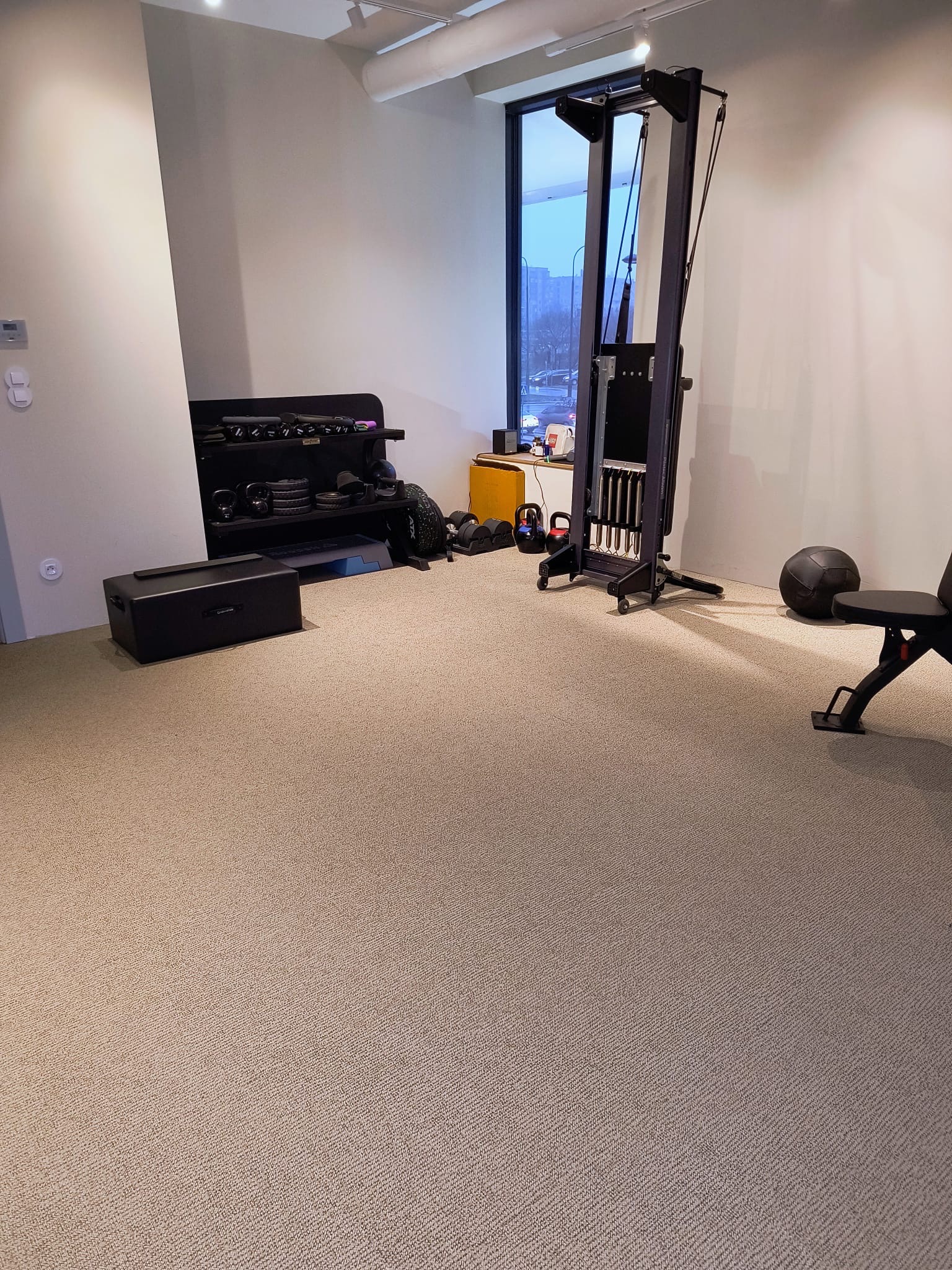 Jasny, beżowy dywan z krótkim włosiem w domowej siłowni z wyposażeniem do ćwiczeń siłowych i akcesoriami.
