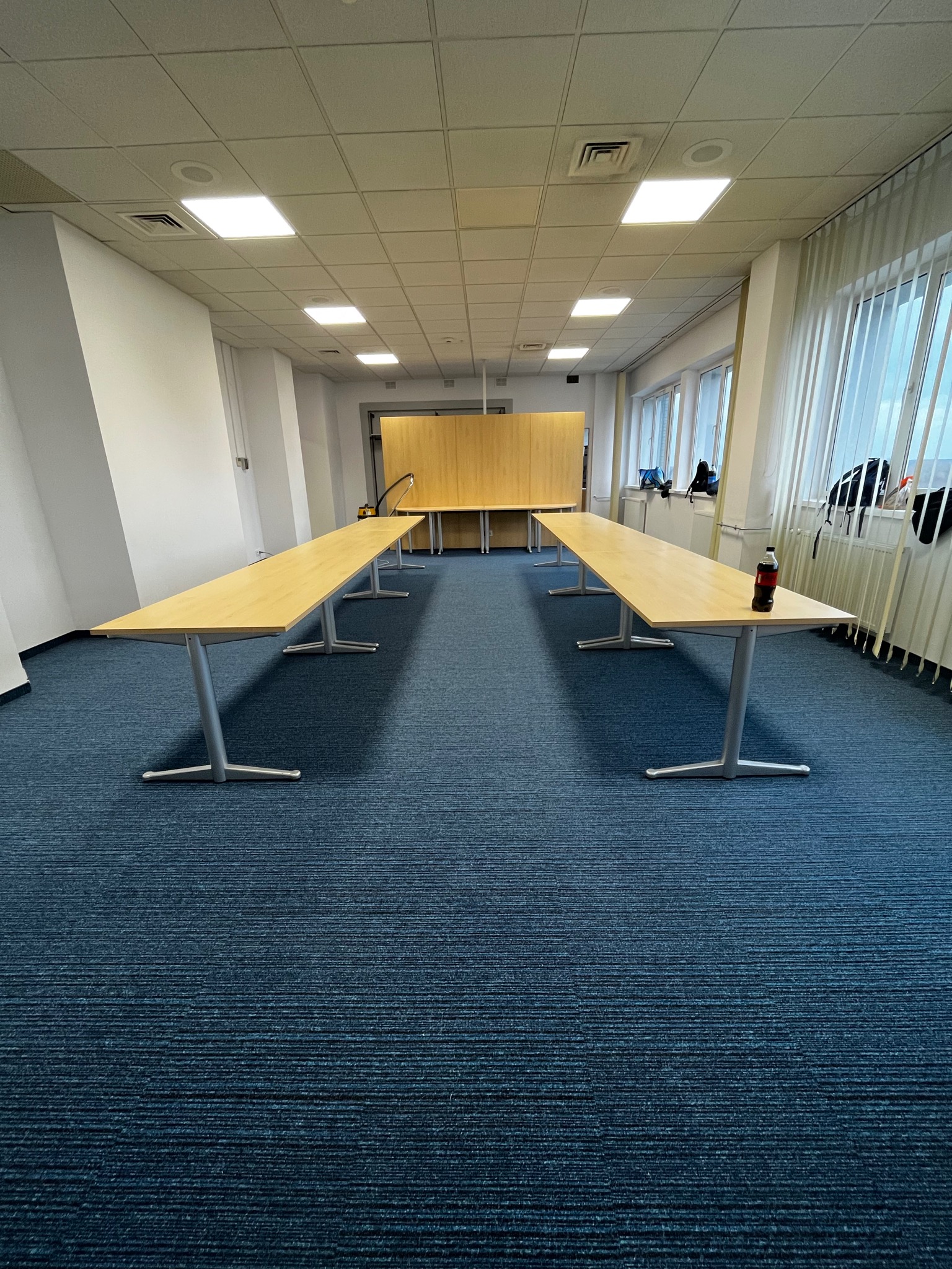 Biuro z długimi stołami na metalowych nogach, pokryte ciemnoniebieską wykładziną dywanową w prążki, z butelką napoju na jednym ze stołów.