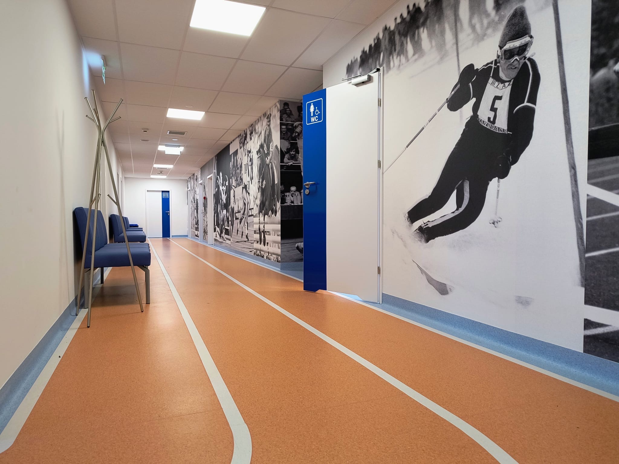 Korytarz z wykładziną imitującą bieżnię sportową, ściany ozdobione czarno-białymi fotografiami sportowców, niebieskie akcenty kolorystyczne.