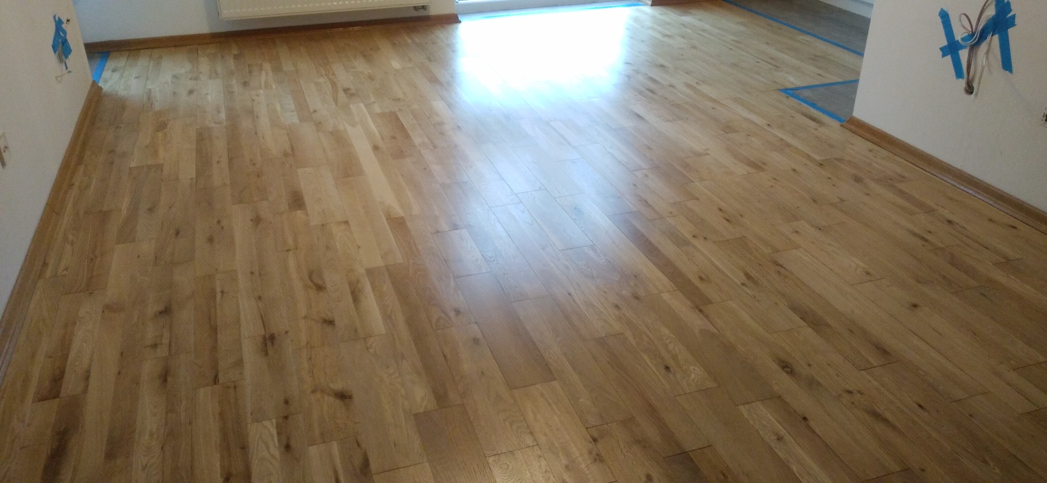 Parkiet dębowy po renowacji, widoczne świeżo położone listwy przypodłogowe, zabezpieczone taśmą malarską przy ścianach.