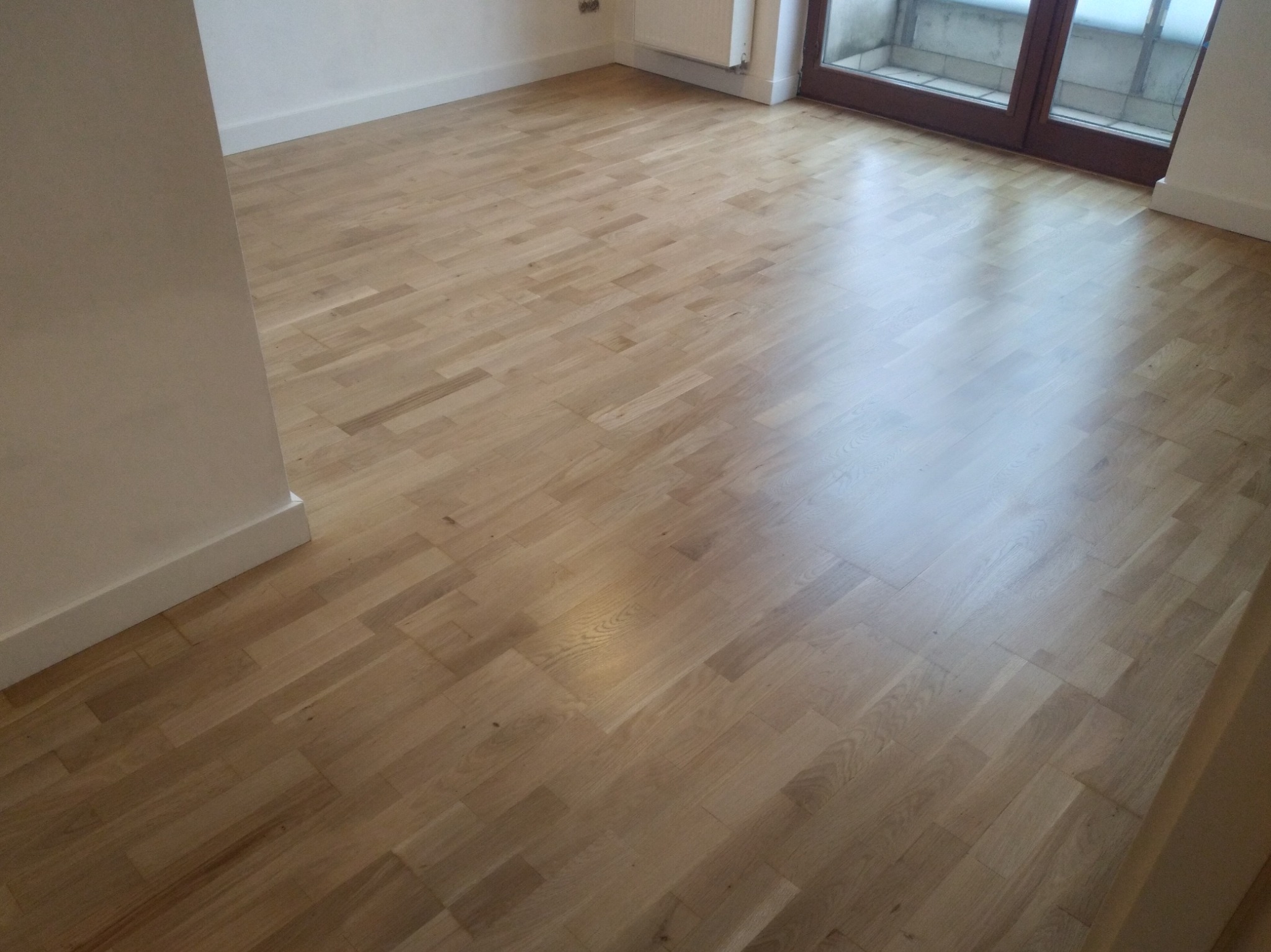 Wycyklinowana, jasna parkietowa podłoga w pustym pokoju z białymi listwami przypodłogowymi i balkonem w tle.