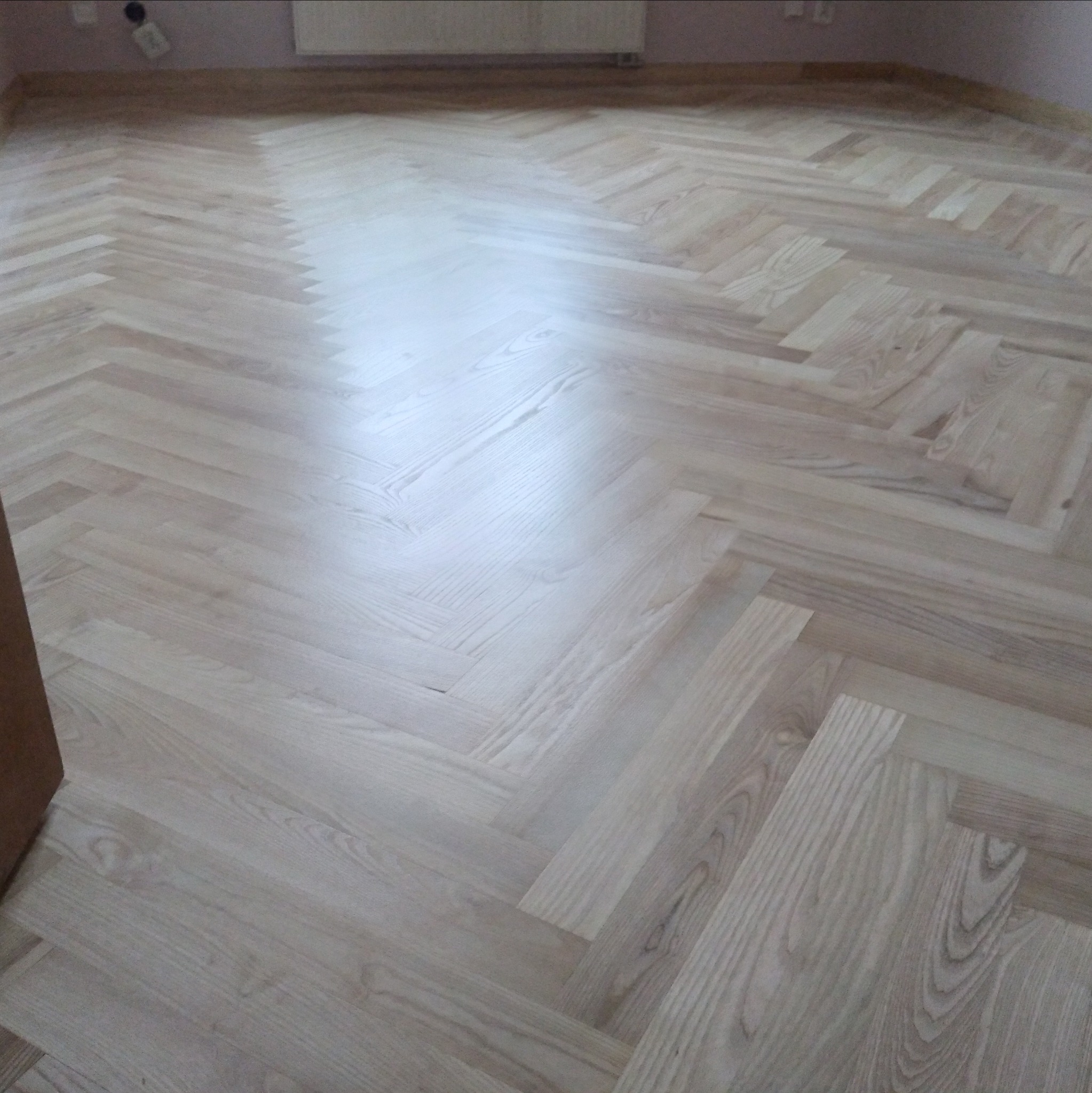 Jasna, odnowiona podłoga parkietowa ułożona we wzór jodełki, z widocznym połyskiem po cyklinowaniu i lakierowaniu, z fragmentem ściany w tle.