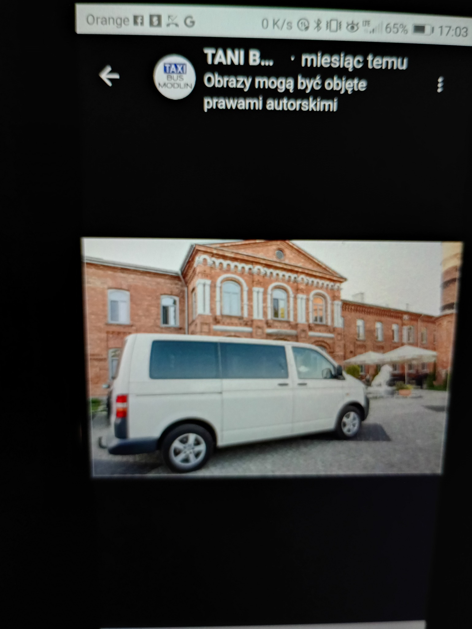 Biały bus typu van na tle ceglanego budynku z arkadami. Pojazd z ciemnymi szybami, zaparkowany na zewnątrz. Widoczne logo Taxi Bus Modlin w lewym górnym rogu.