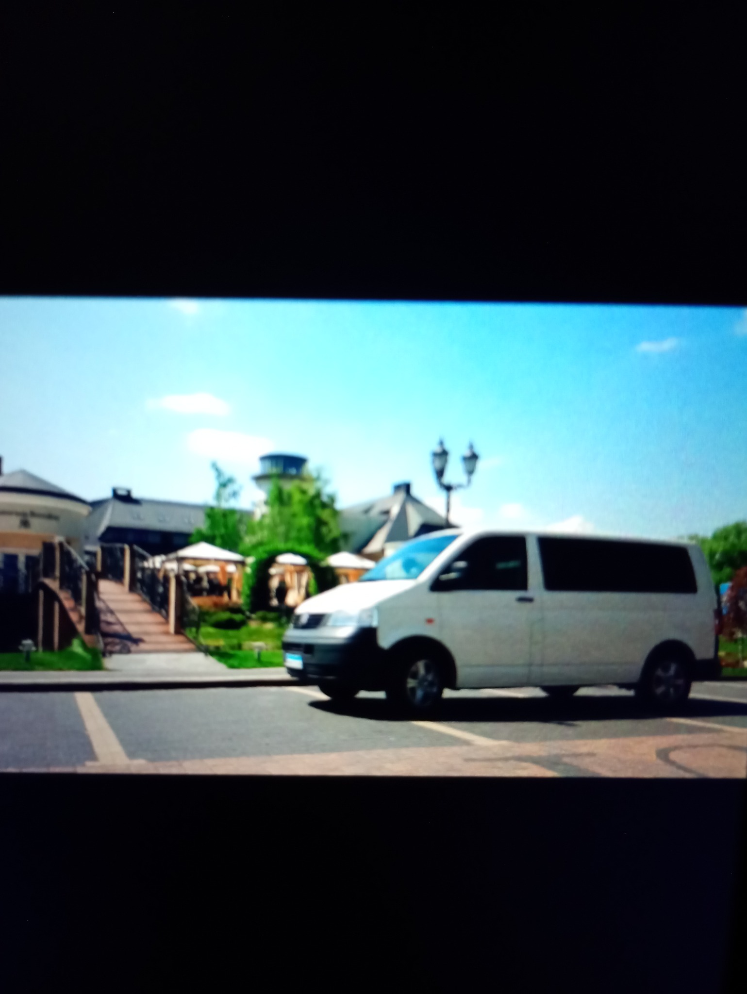 Biały bus na tle eleganckiego budynku z ogrodem i mostkiem. Usługa transportowa w otoczeniu zieleni i architektury.