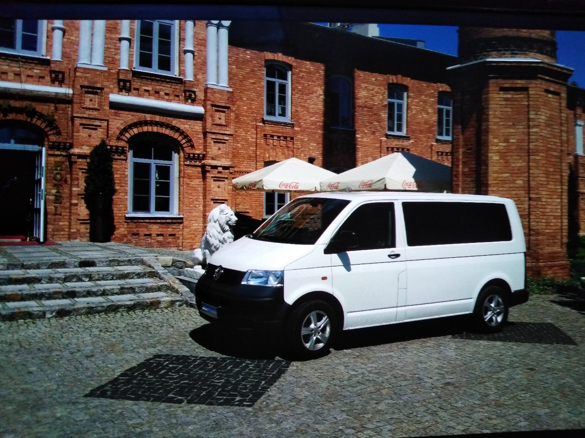 Biały bus VW Transporter T5 na brukowanym dziedzińcu przed ceglanym budynkiem z kolumnami i rzeźbą lwa. Dwa parasole Coca-Cola w tle.