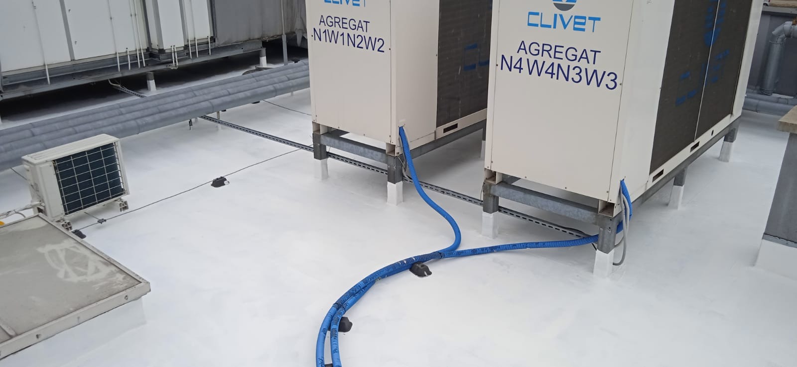 Dwa agregaty klimatyzacyjne marki Clivet na dachu budynku, z widocznymi niebieskimi przewodami i instalacją elektryczną w korytkach, całość na białym, izolowanym dachu.
