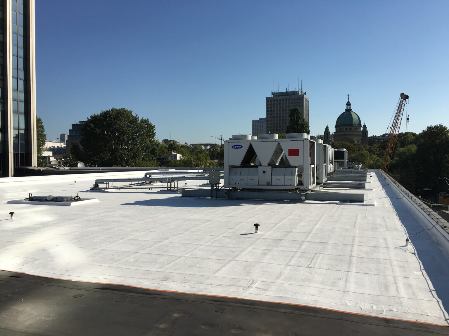 Biały dach budynku z zainstalowanymi jednostkami klimatyzacji Carrier, widok na panoramę miasta z kopułą kościoła w tle.