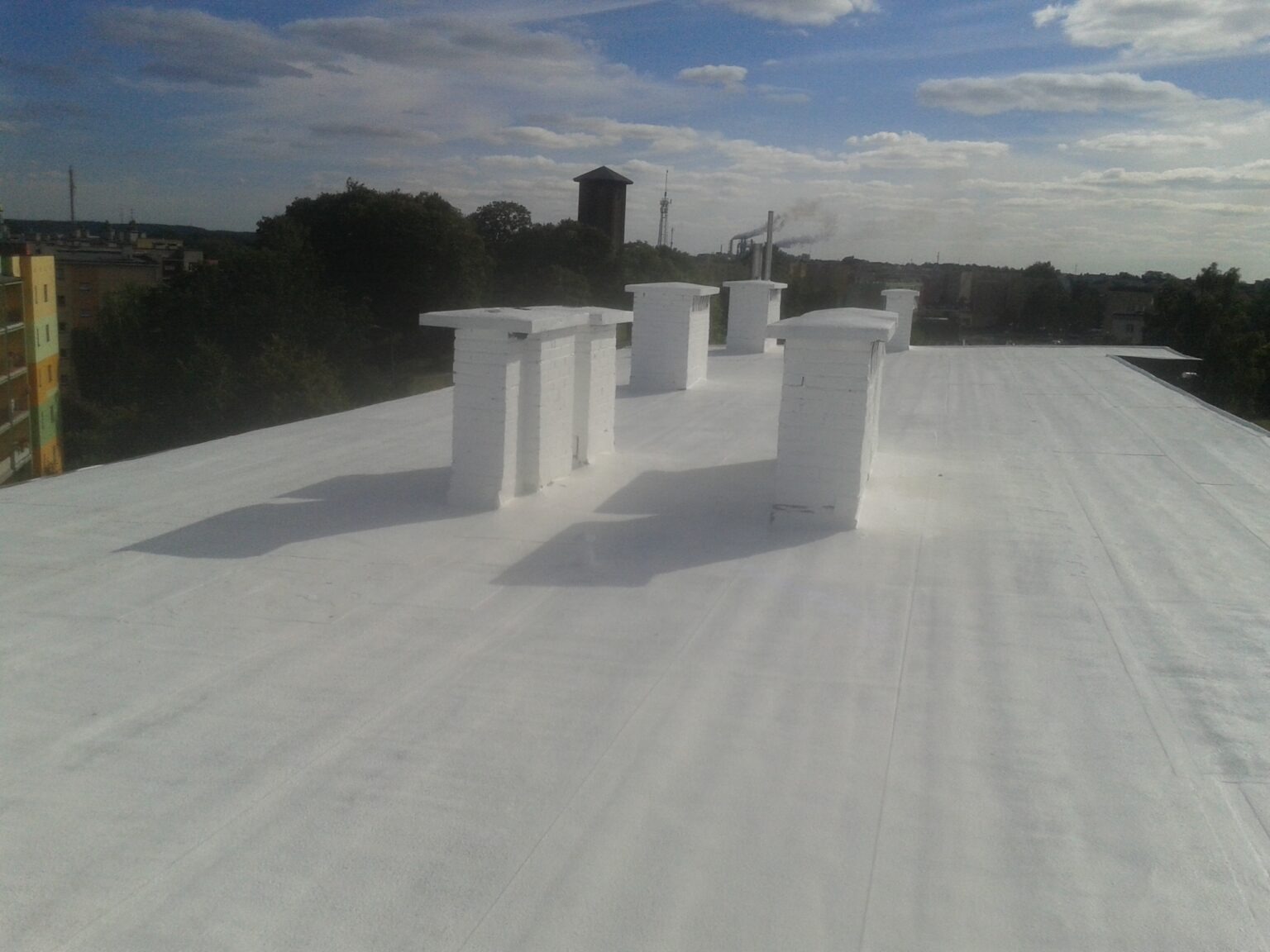 Biały, płaski dach po renowacji z kominami pomalowanymi na biało, widok na miasto z kominem fabrycznym w tle, lekko pochmurne niebo.