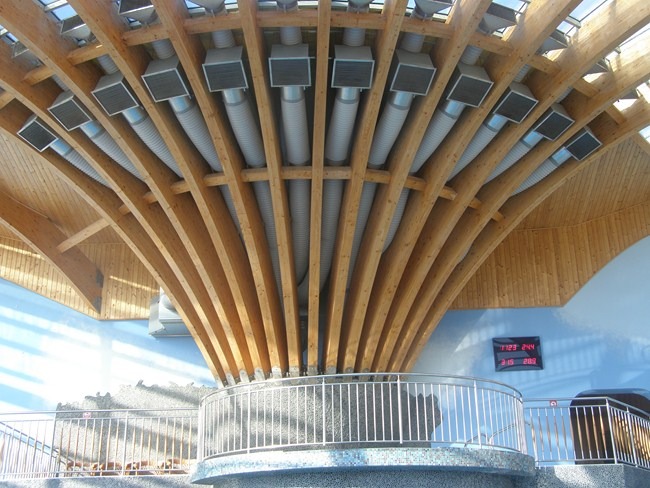 Wnętrze budynku z widoczną drewnianą konstrukcją dachu i systemem wentylacji z licznymi kanałami wentylacyjnymi, zakończonymi kratkami, oraz elektronicznym wyświetlaczem czasu i temperatury...