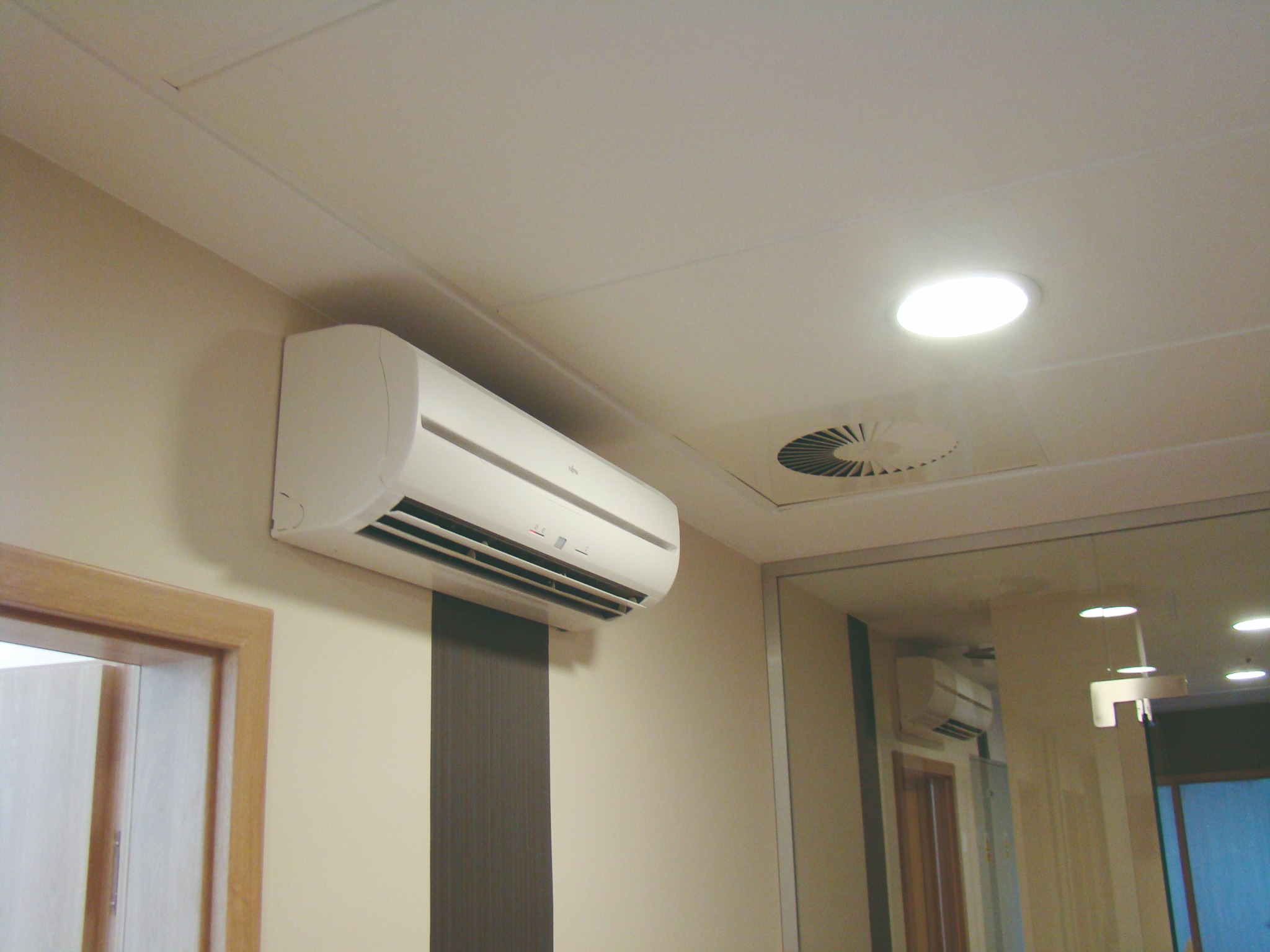 Biały klimatyzator ścienny zamontowany w jasnym pomieszczeniu, widoczny również okrągły nawiewnik wentylacyjny oraz odbicie klimatyzatora w lustrze.