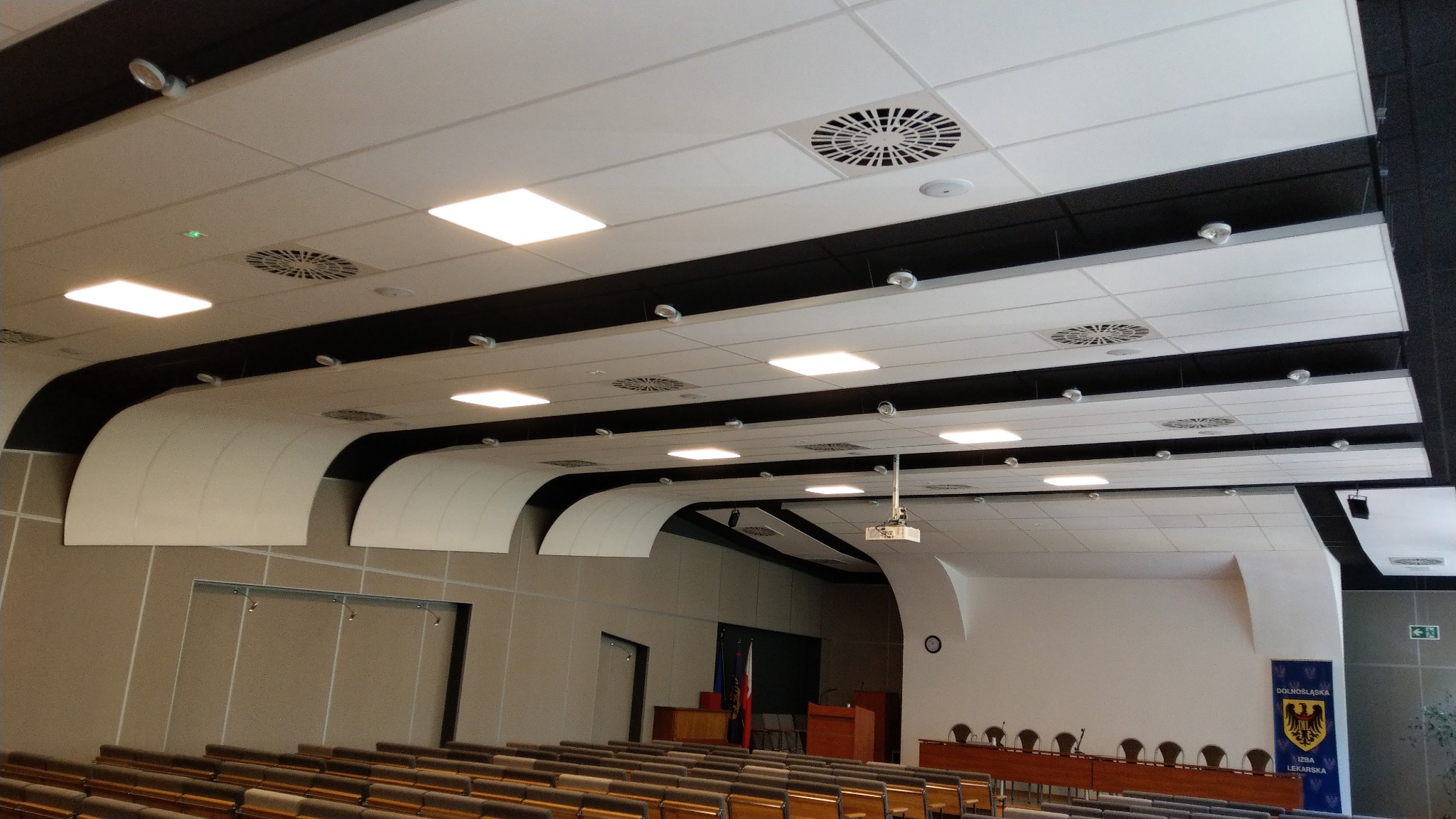 Sufit podwieszany w dużej sali konferencyjnej z widocznymi nawiewnikami wentylacyjnymi, oświetleniem panelowym LED i elementami dekoracyjnymi w kształcie łuków.