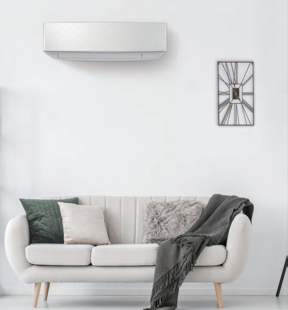 Biały klimatyzator ścienny zamontowany nad kremową sofą z poduszkami w odcieniach szarości i zieleni, na białej ścianie wisi ozdobny zegar.