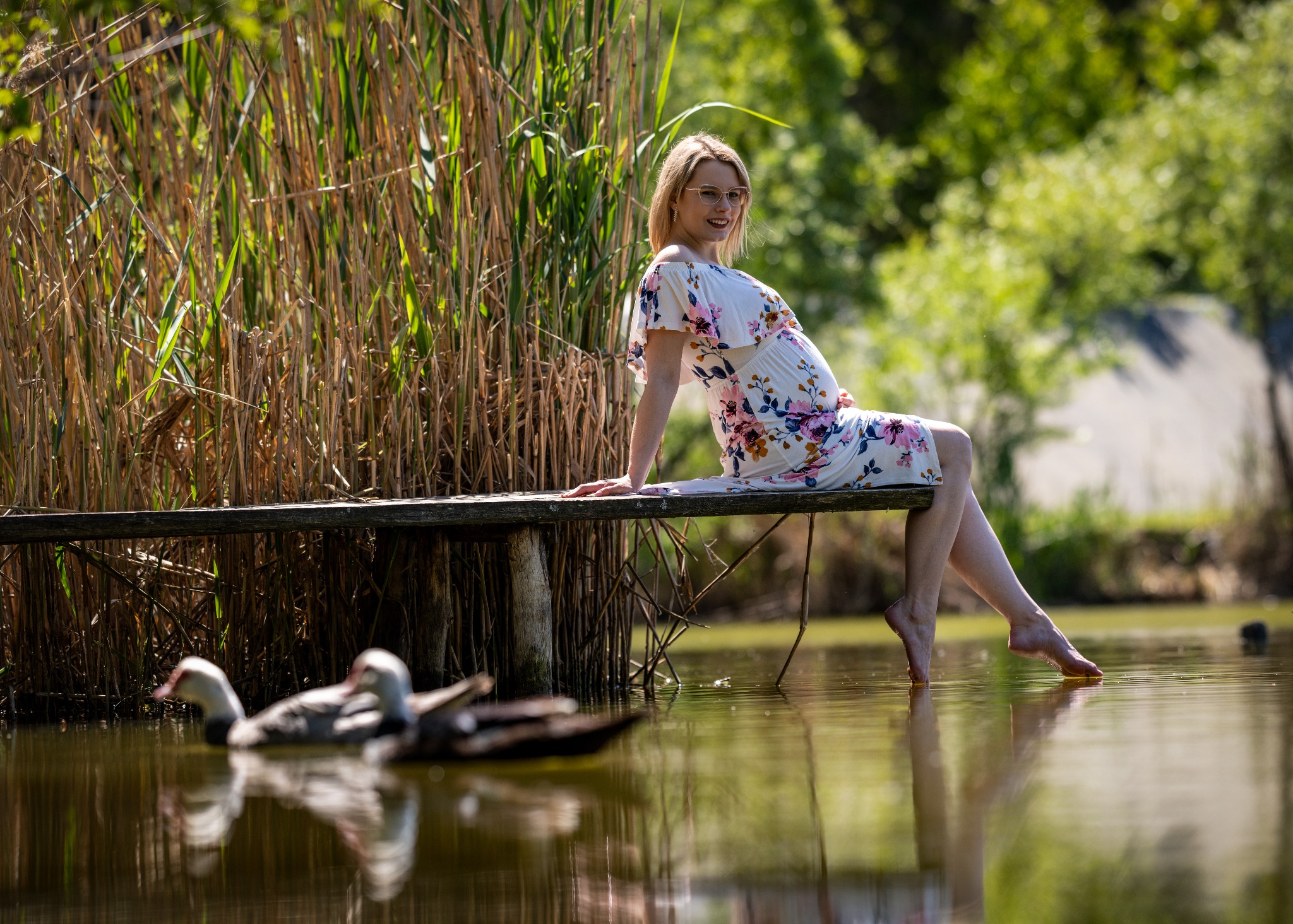 Uśmiechnięta, ciężarna blondynka w kwiecistej sukience siedzi na drewnianym pomoście nad wodą, obok szuwarów, z kaczkami w tle.