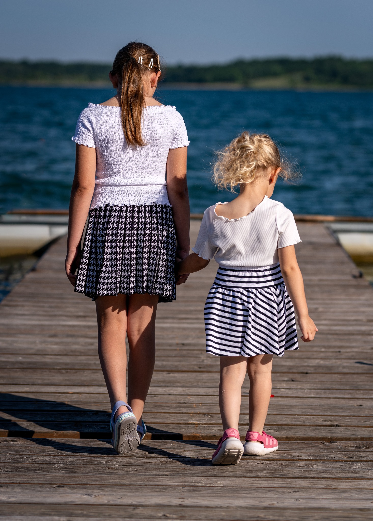 Dwie dziewczynki, starsza i młodsza, idą trzymając się za ręce po drewnianym pomoście w kierunku jeziora w słoneczny dzień. Starsza ma spódnicę w pepitkę, a młodsza w paski.