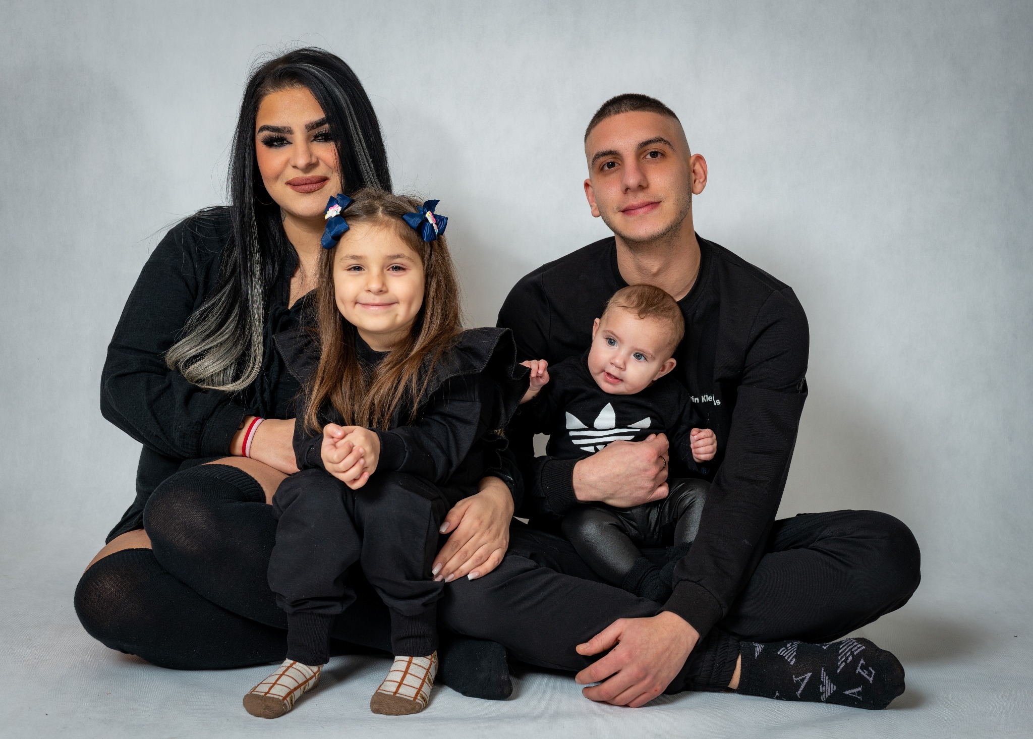 Portret rodzinny: matka z pasemkami, ojciec trzymający niemowlę w ubranku Adidas, córka z kokardami, wszyscy ubrani na czarno, na jasnoszarym tle.