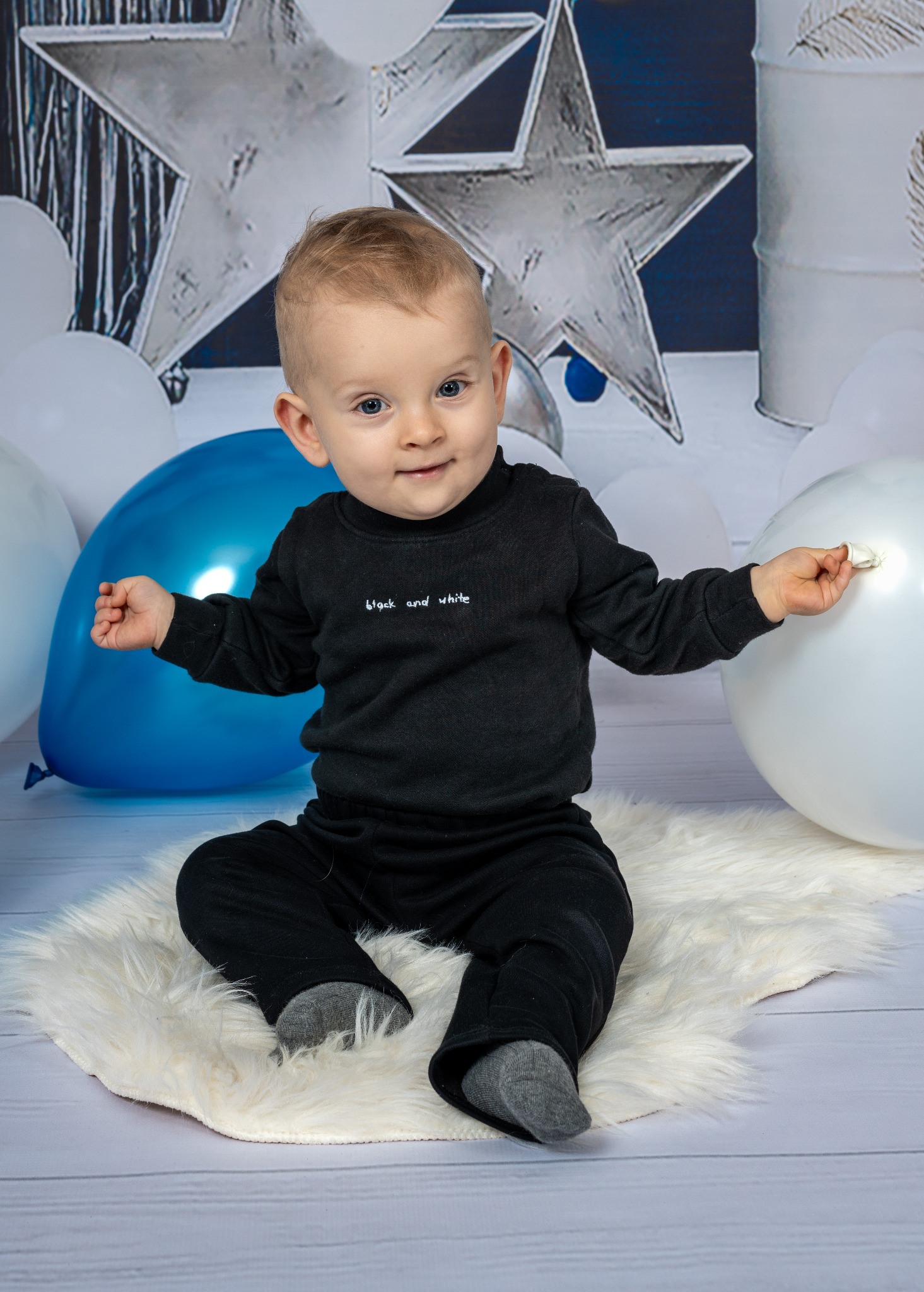 Uśmiechnięty roczny chłopiec w czarnym ubranku siedzi na białym futrzanym dywaniku, otoczony białymi i niebieskimi balonami, na tle dekoracji z gwiazd i beczek.