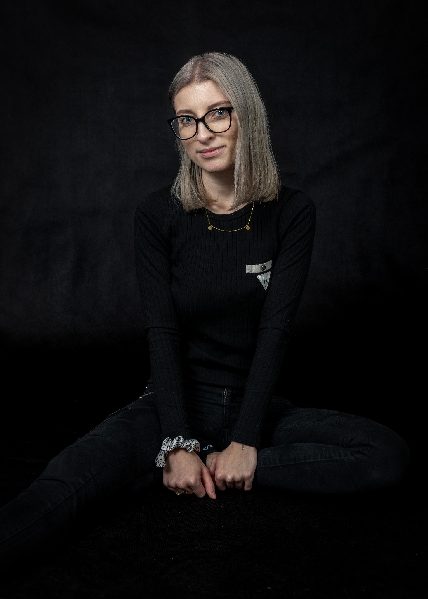 Portret kobiety w okularach, siedzącej na czarnym tle, ubrana w czarny sweter i dżinsy, z delikatnym uśmiechem i platynowymi włosami.