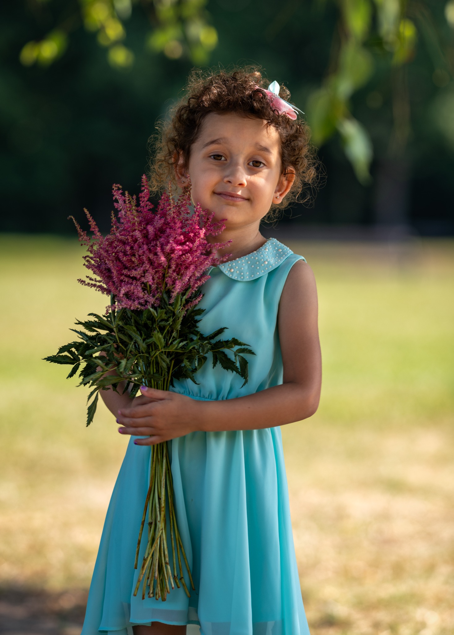 Uśmiechnięta dziewczynka w błękitnej sukience trzyma bukiet różowych kwiatów na tle zielonego parku.