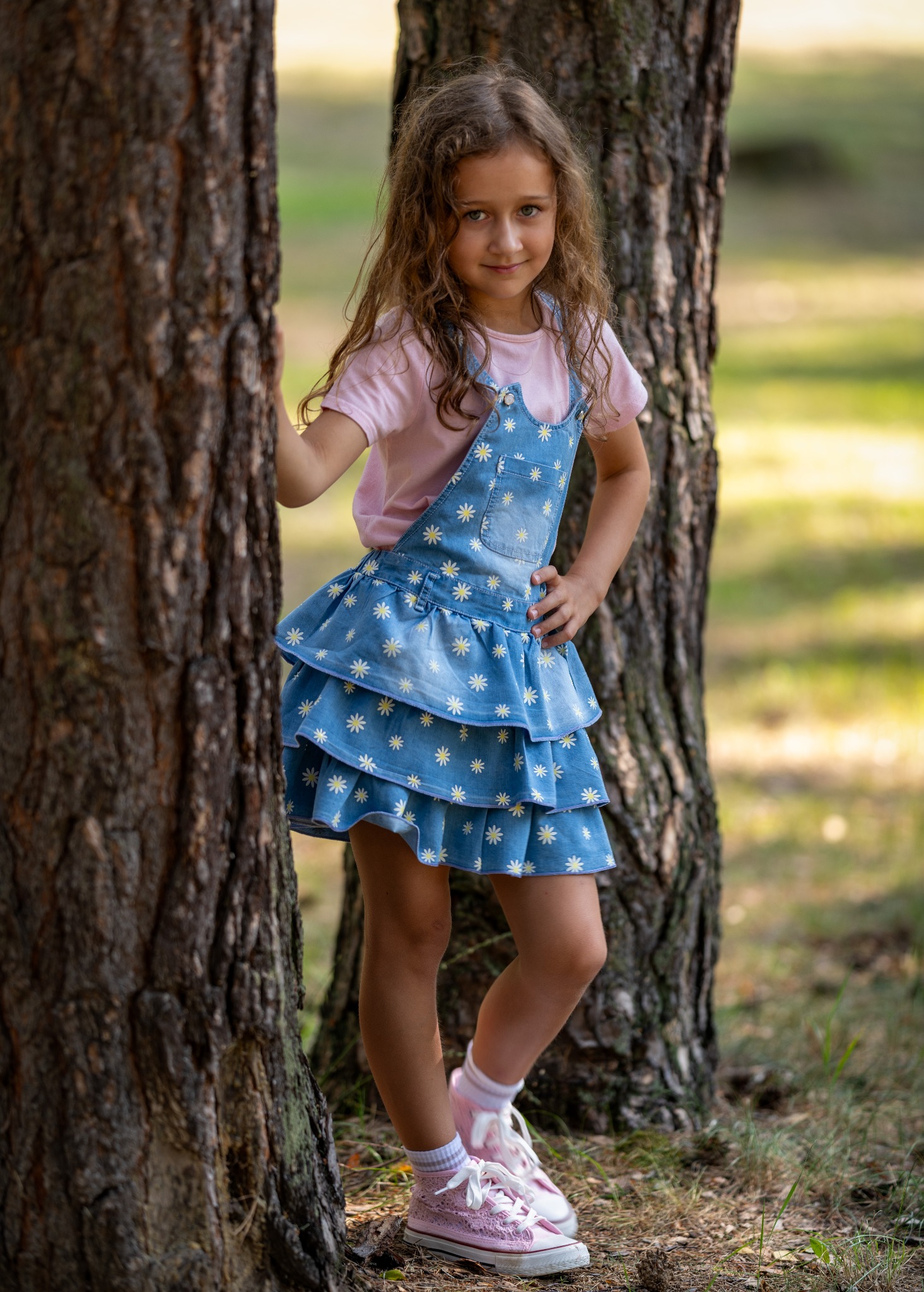Portret uśmiechniętej dziewczynki w dżinsowej sukience w kwiatki, stojącej między dwoma drzewami w lesie, z ręką na biodrze i różowymi brokatowymi butami.