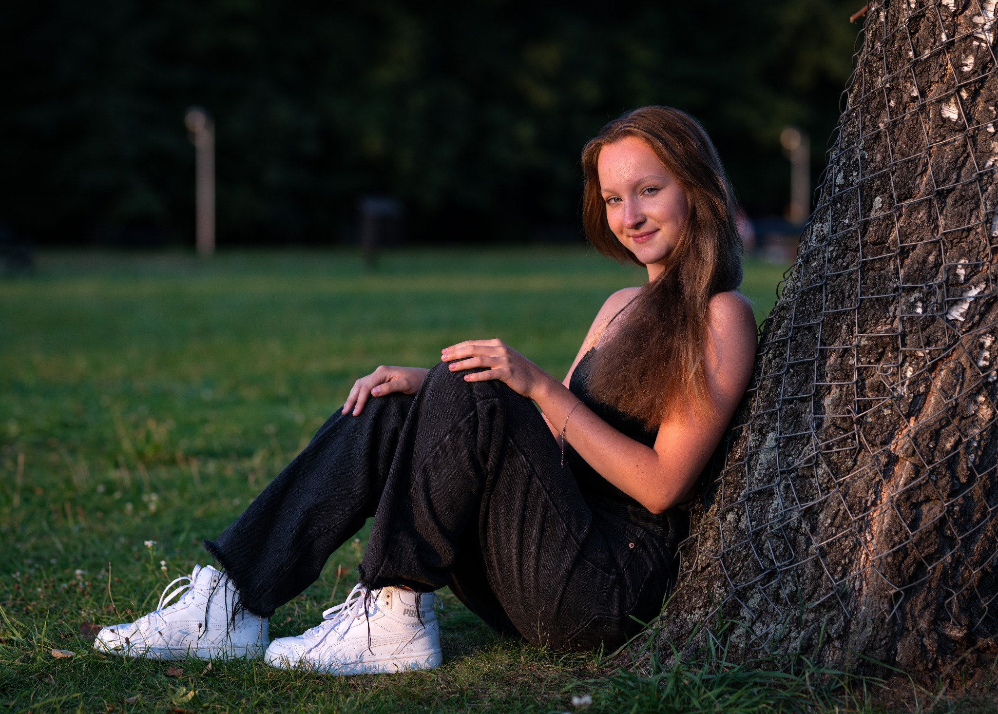 Młoda kobieta z długimi, rudymi włosami, ubrana w czarny top i szerokie jeansy, opiera się plecami o drzewo w parku, uśmiechając się do kamery. Na nogach ma białe sportowe buty Puma.
