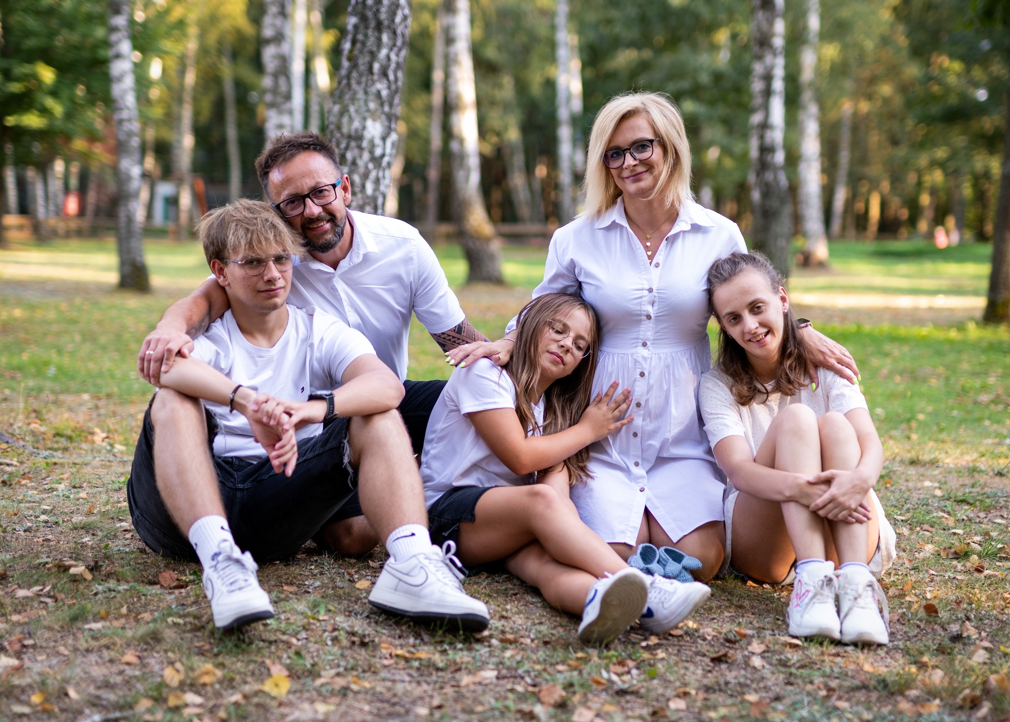 Rodzinna sesja w plenerze: rodzice i troje dzieci siedzą na ziemi, w tle brzozowy zagajnik, naturalne światło.