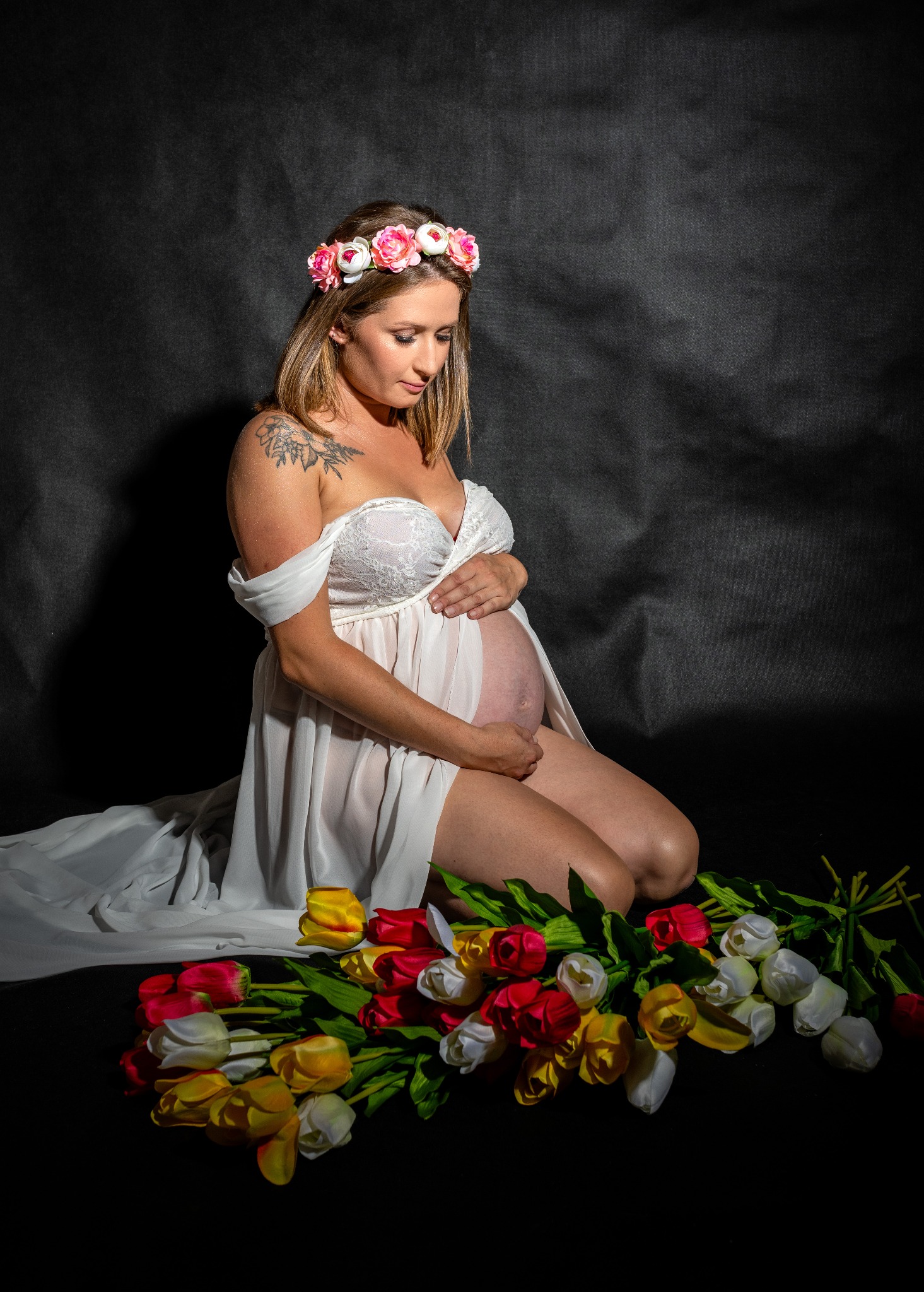 Kobieta w ciąży, w białej sukni i wianku z kwiatów na głowie, klęczy na czarnym tle, trzymając ręce na brzuchu, obok leżą kolorowe tulipany.