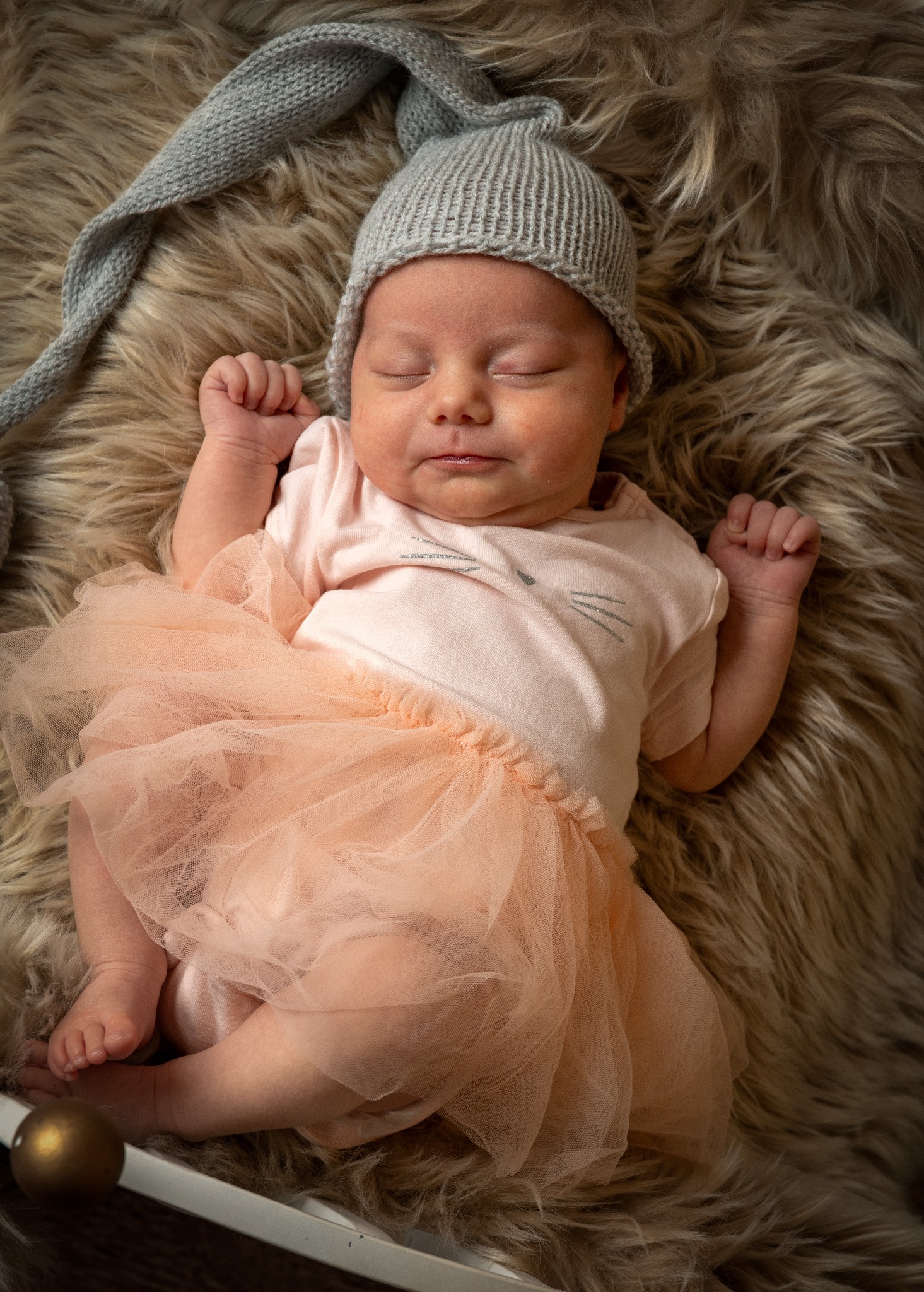 Śpiąca noworodka w szarej czapce z pomponem i różowej tiulowej spódniczce, leżąca na futrzanym tle, z widoczną kulą dekoracyjną w rogu kadru.