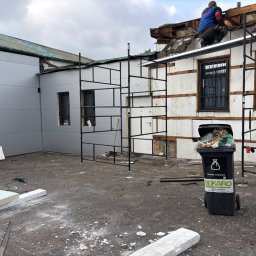 DEKS Jarosław Starak - Pracownik na rusztowaniu demontuje fragment dachu budynku z widocznymi panelami i oknami z kratami; na pierwszym planie śmieci i narzędzia.