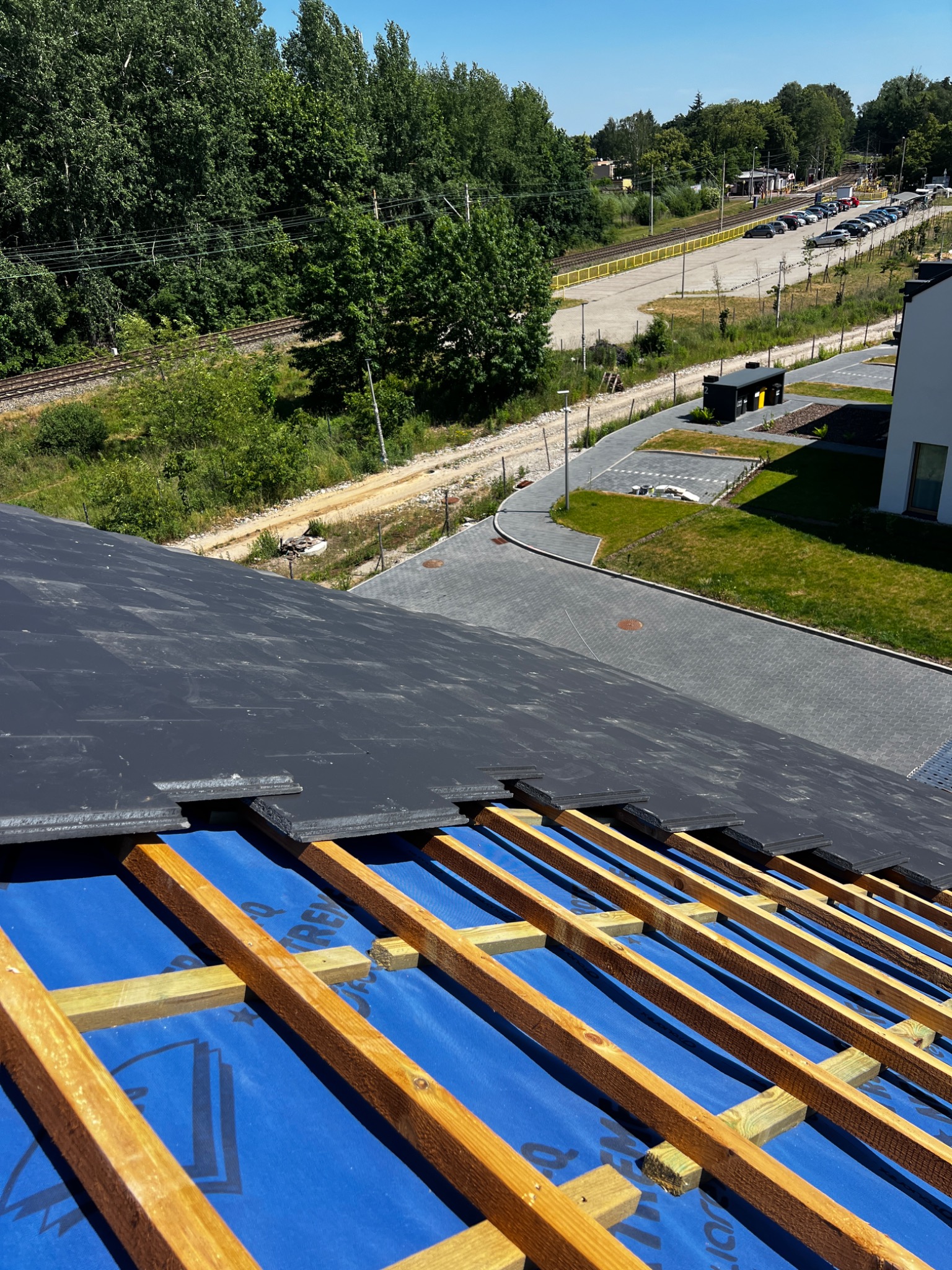 Fragment dachu w trakcie układania czarnej dachówki na niebieskiej membranie, widoczne drewniane łaty, w tle budynek, zieleń i tory kolejowe.