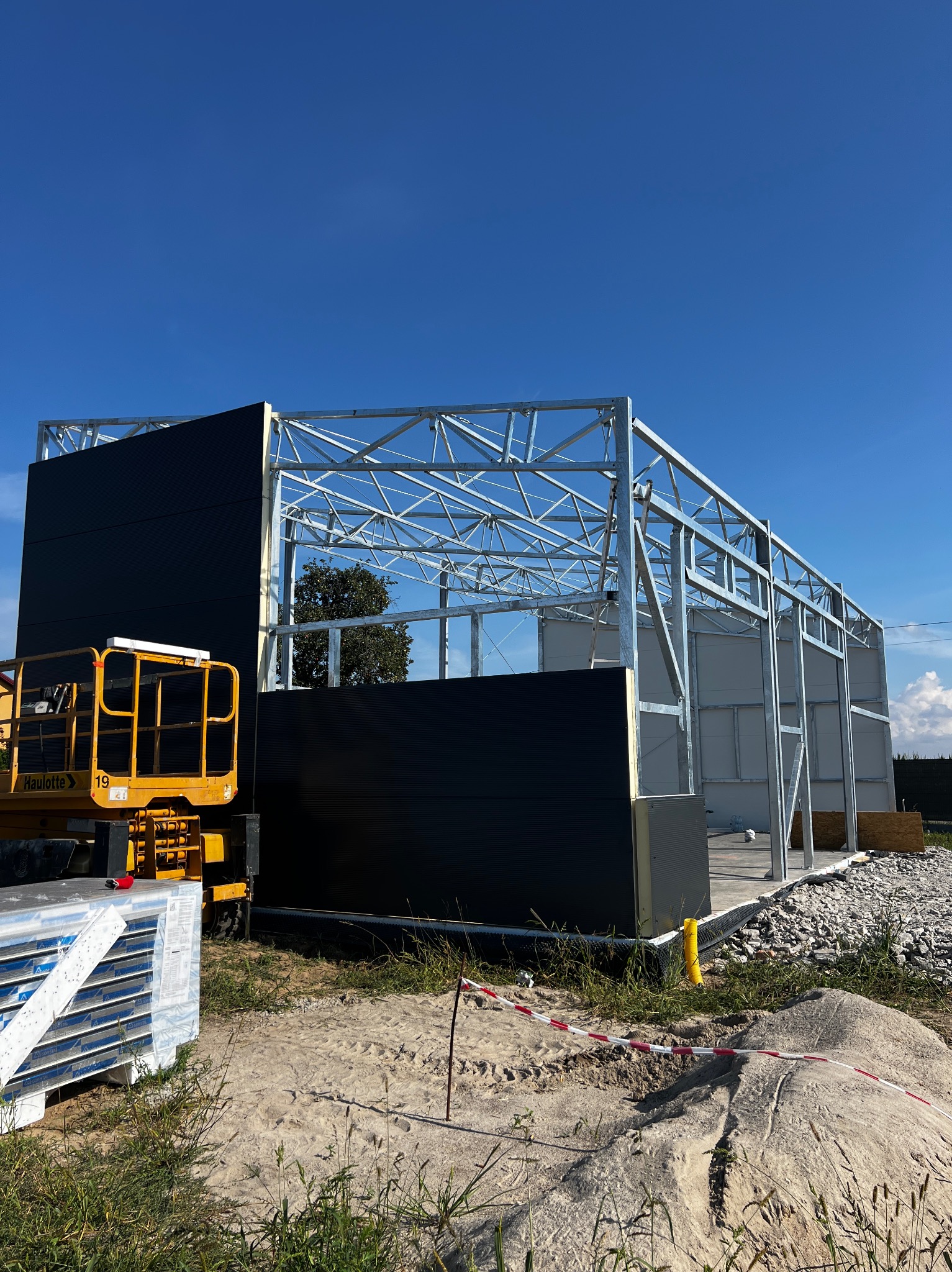 Budowa hali: stalowa konstrukcja szkieletowa z zamontowanymi fragmentami czarnych płyt warstwowych, żółty podnośnik Haulotte z boku, paleta płyt warstwowych, piasek i trawa w tle, błękitne niebo.