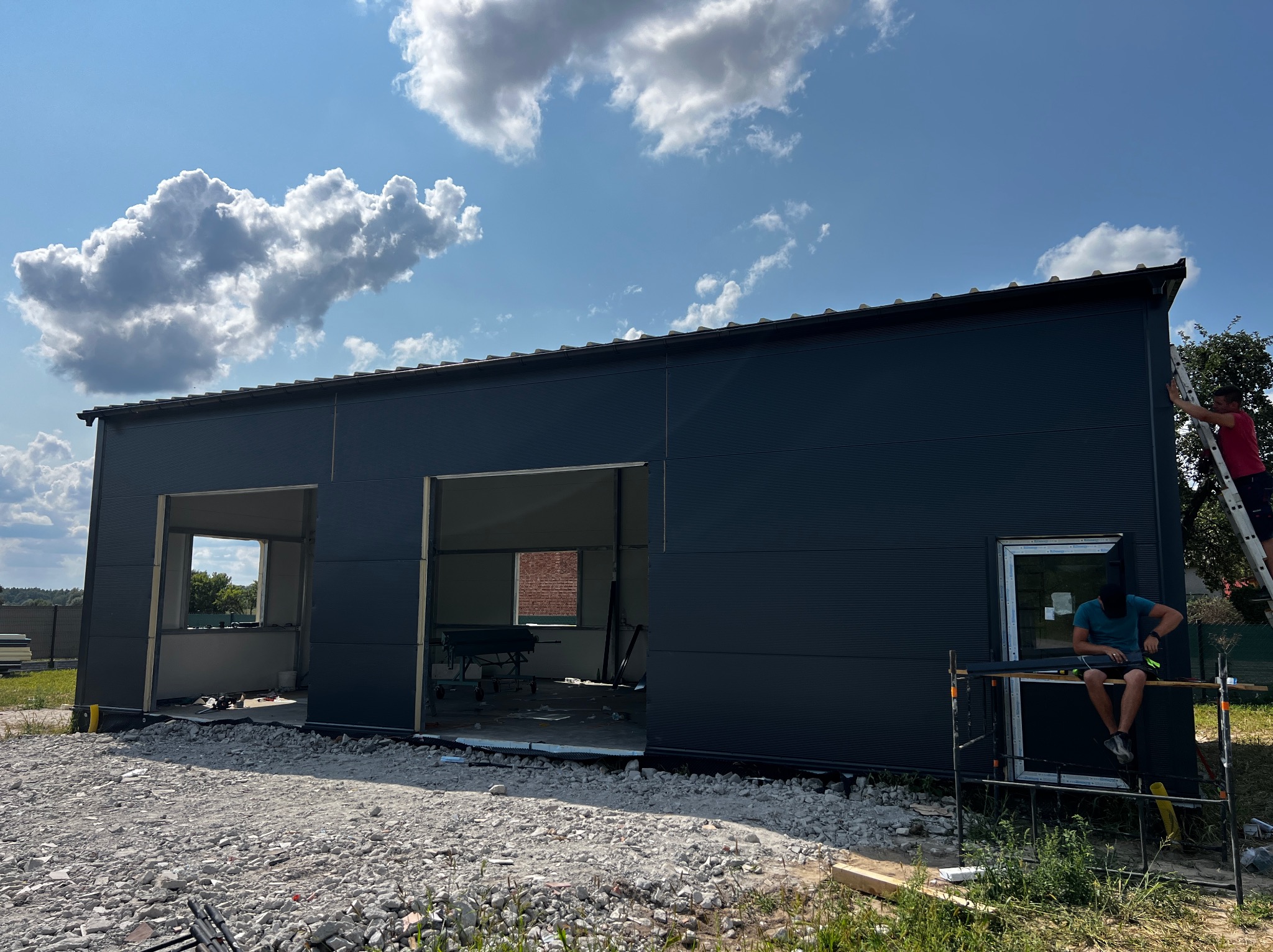 Wznoszona hala z płyt warstwowych w trakcie montażu, widoczne otwory na bramy, pracownicy na rusztowaniu i drabinie, teren budowy z gruzem i materiałami budowlanymi, błękitne niebo z chmurami.