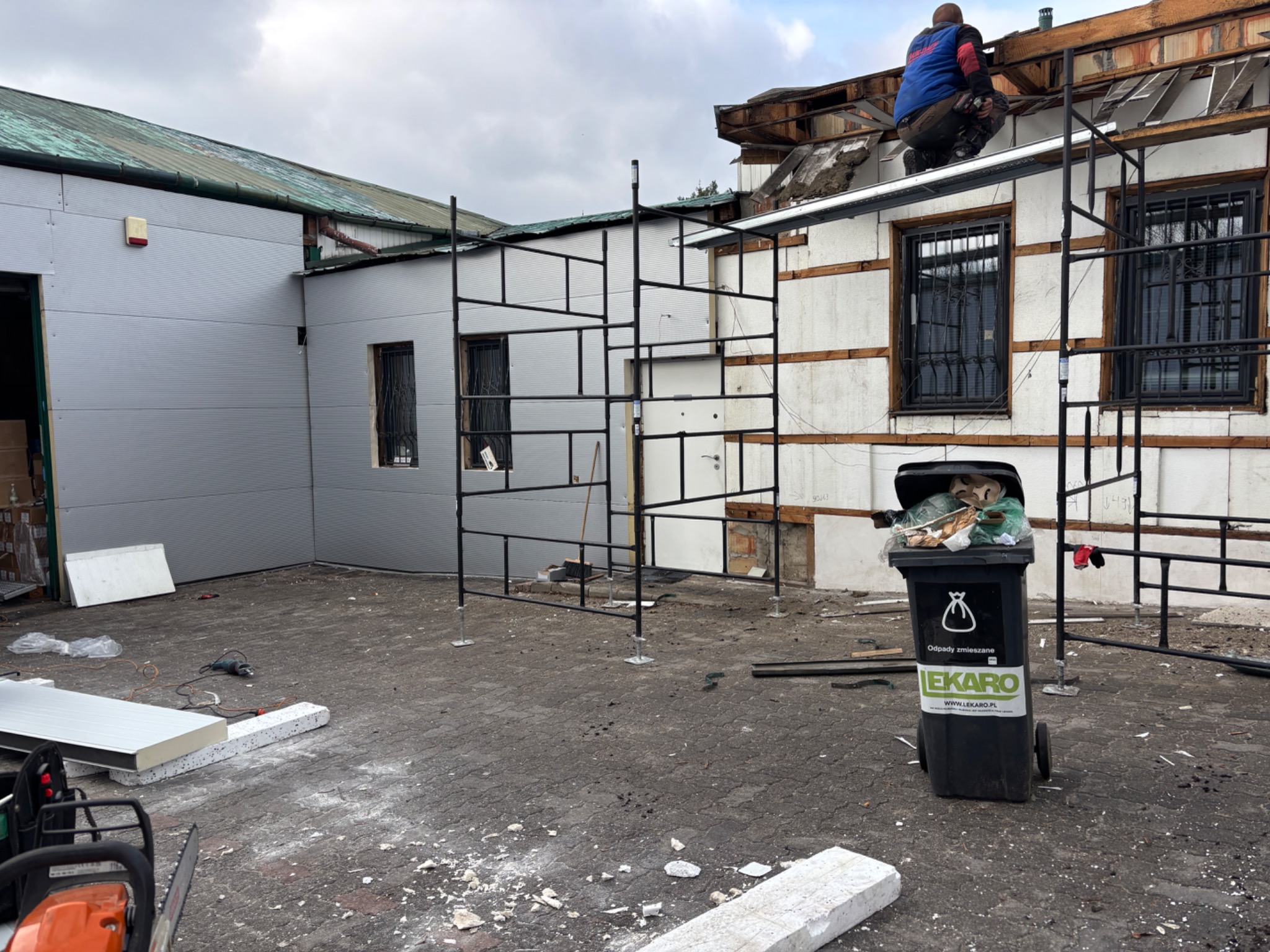 Pracownik na rusztowaniu demontuje fragment dachu budynku z widocznymi panelami i oknami z kratami; na pierwszym planie śmieci i narzędzia.