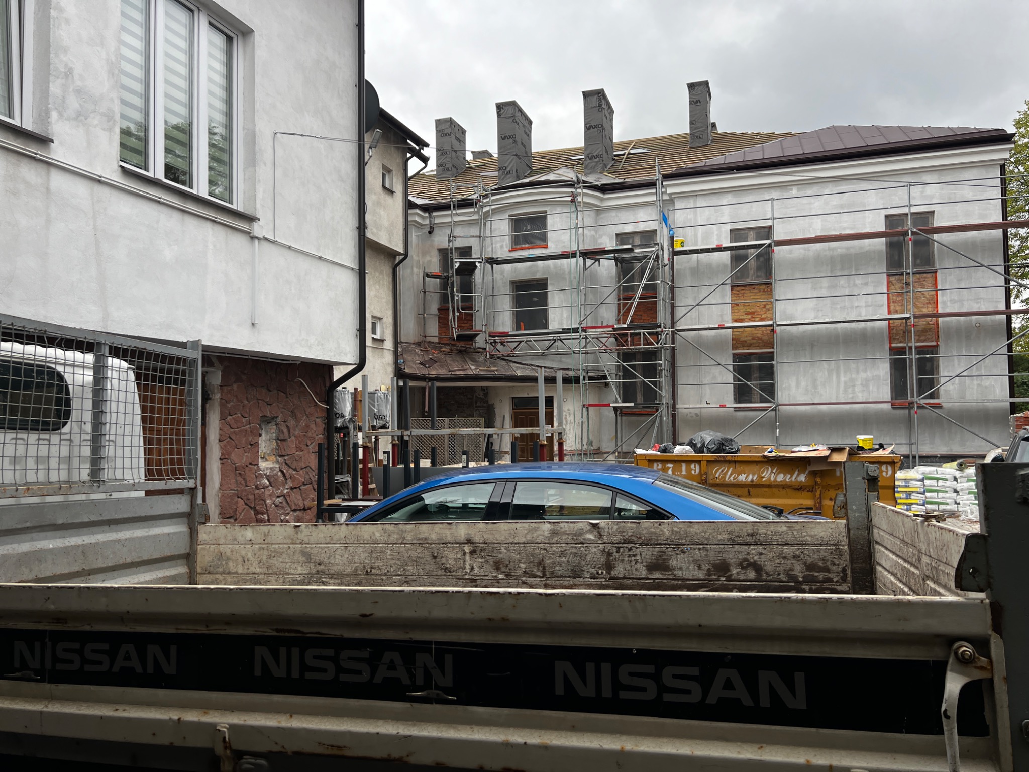 Budynek w trakcie remontu dachu, widoczny z perspektywy skrzyni samochodu dostawczego, rusztowania, nowe kominy zabezpieczone folią, worki z materiałami budowlanymi obok kontenera na odpady.