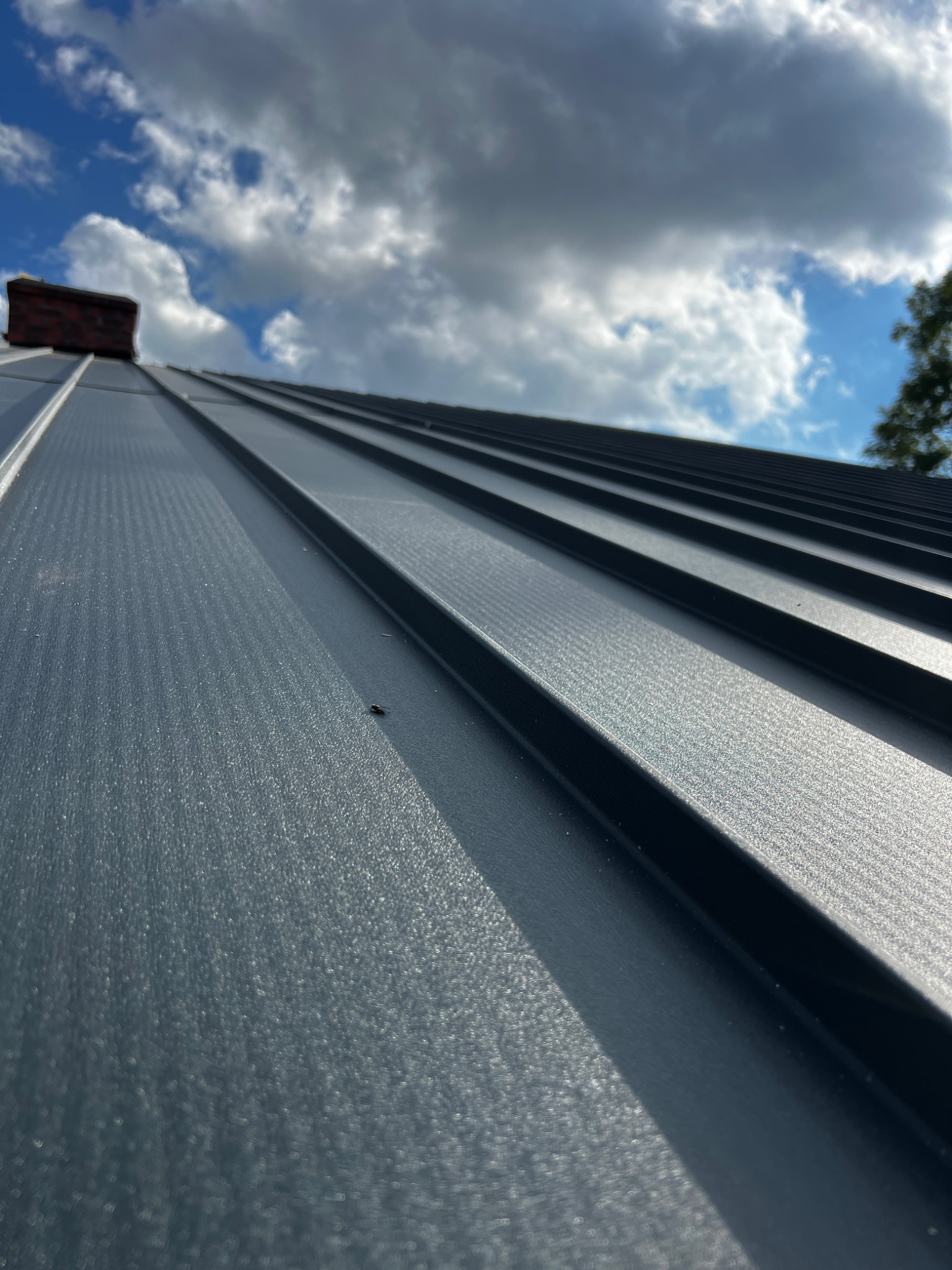 Nowoczesny, ciemnoszary dach z blachy trapezowej, widok z dołu, z kominem w tle i fragmentem błękitnego nieba z obłokami.