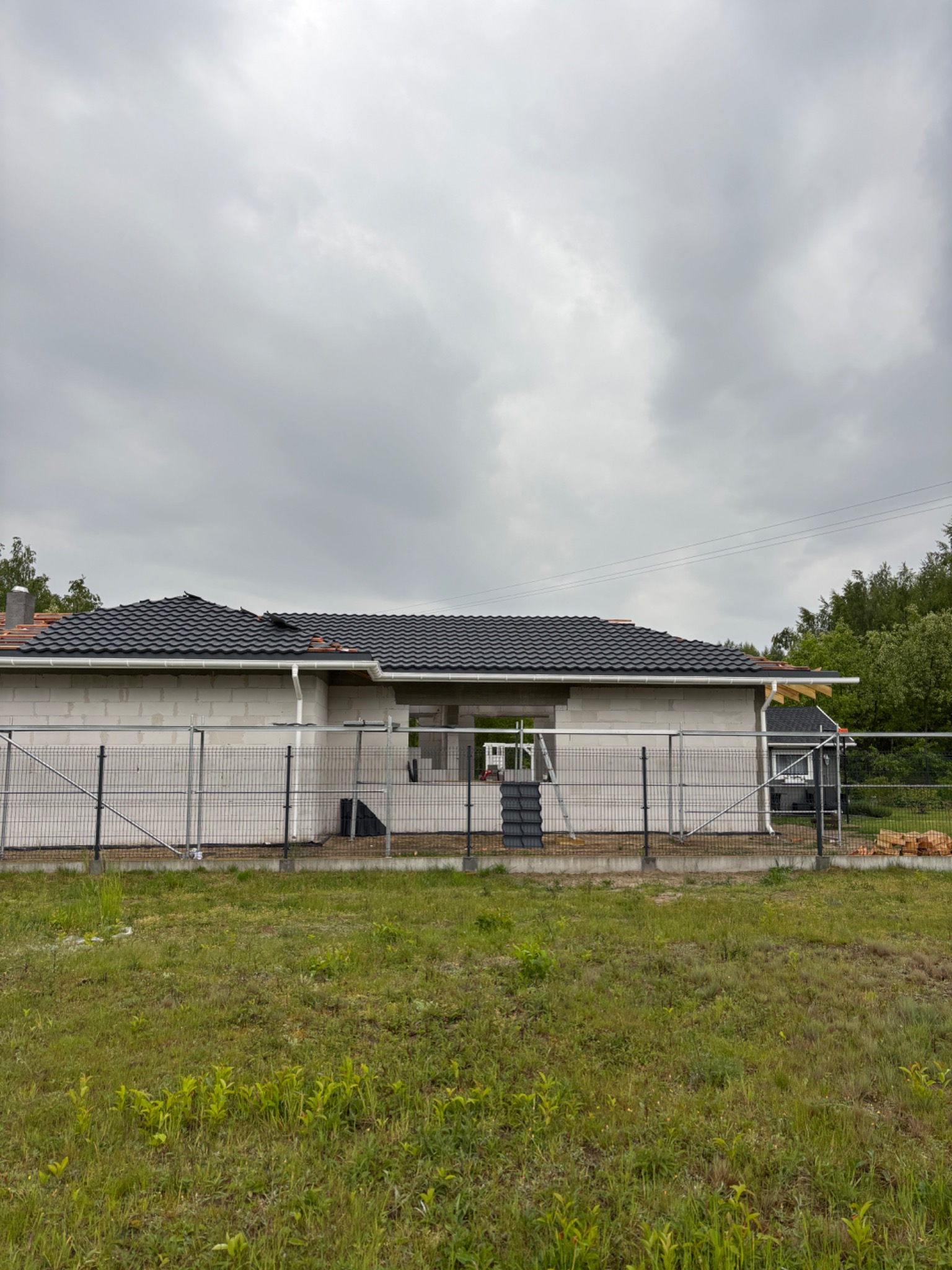 Dom w trakcie krycia dachu czarną dachówką. Widoczne rusztowania i materiały budowlane. Prace dekarskie na etapie układania pokrycia.