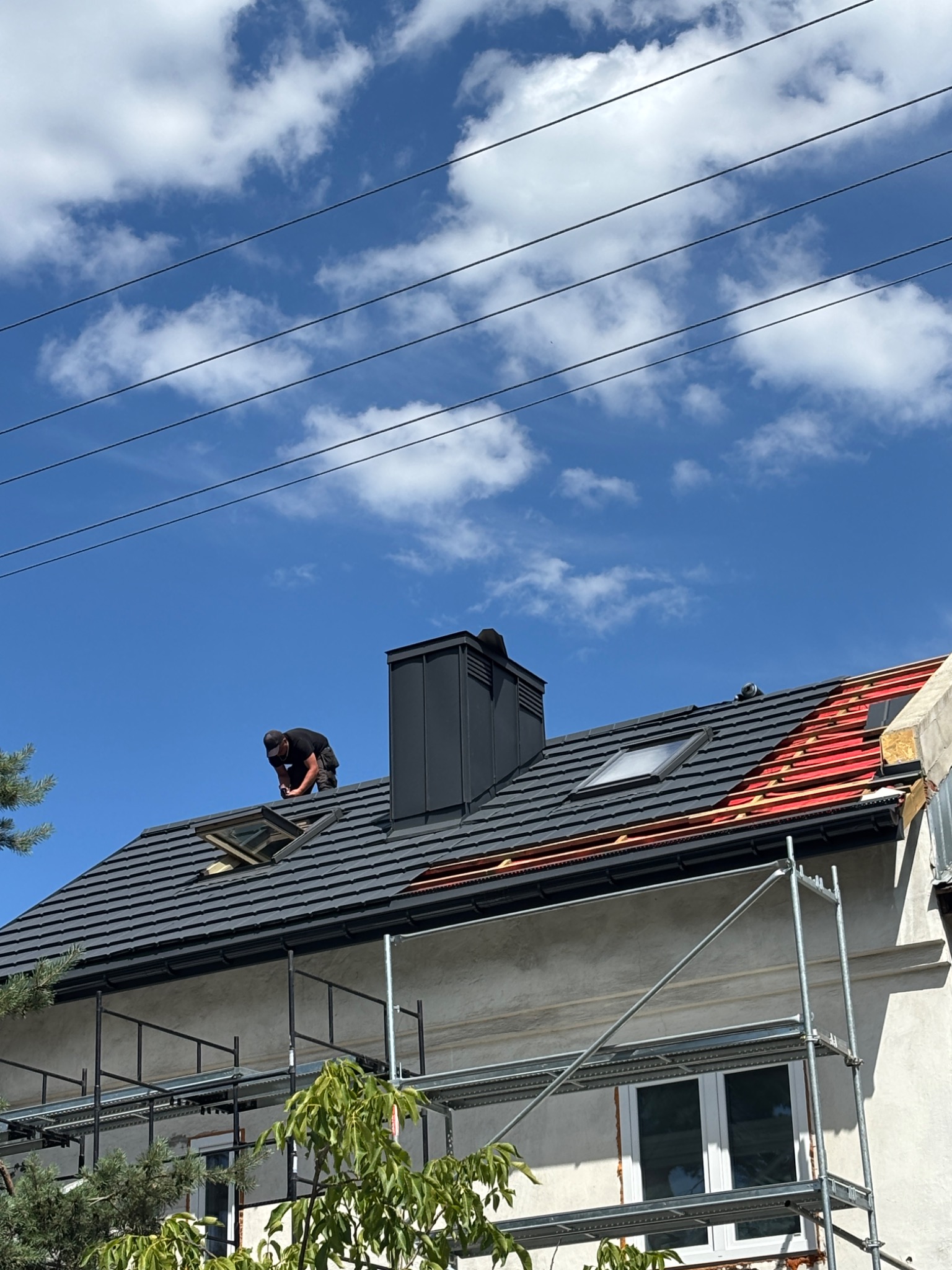 Praca na dachu domu: dekarz montuje okno dachowe na tle błękitnego nieba z chmurami. Widoczne rusztowania i fragmenty dachu z czerwoną membraną.