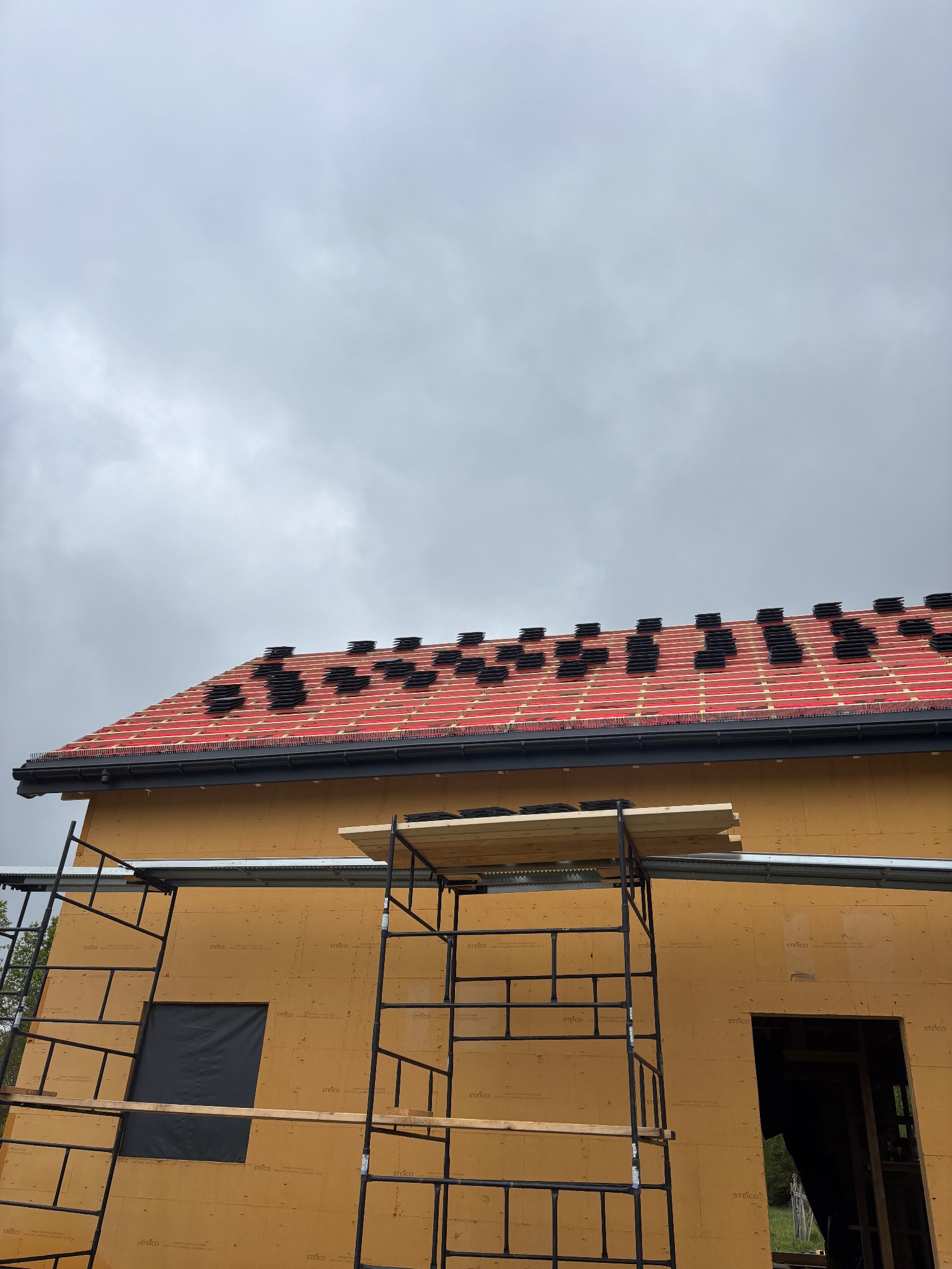 Dach w trakcie krycia: czerwona membrana i czarne dachówki ułożone rzędami. Widoczne rusztowanie i fragment ściany budynku z płyt OSB. Pochmurne niebo w tle.