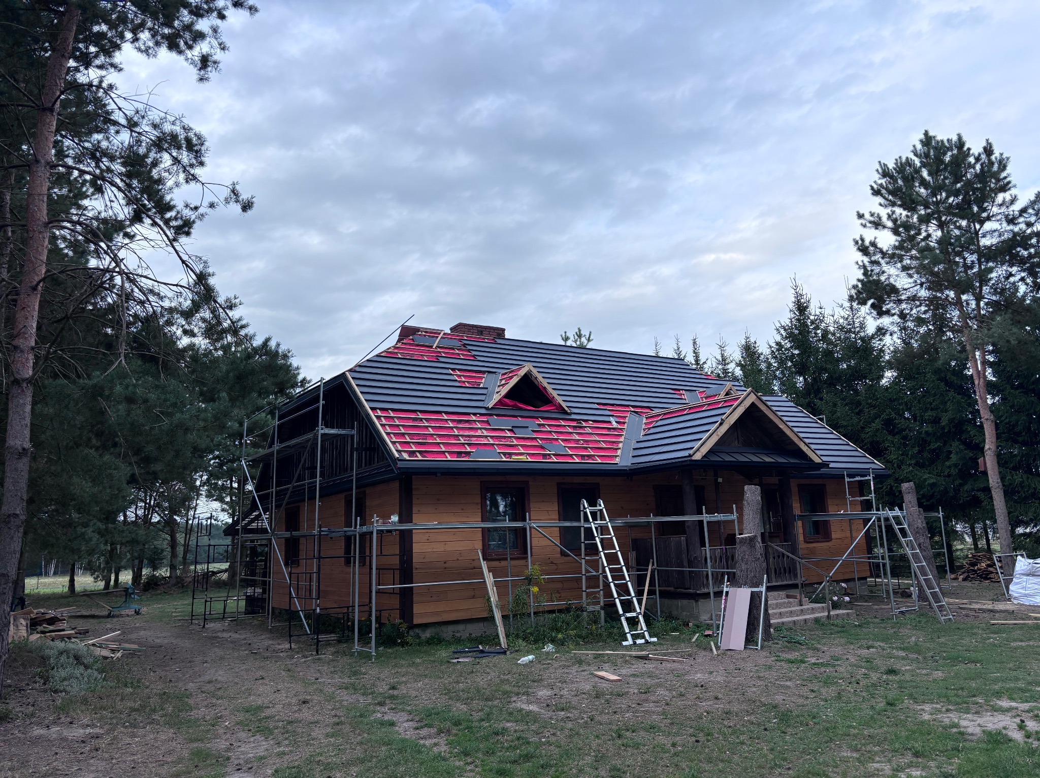 Dom z częściowo położonym, grafitowym dachem, widoczne rusztowania i elementy konstrukcji dachu. Prace dekarskie w toku, otoczenie leśne.