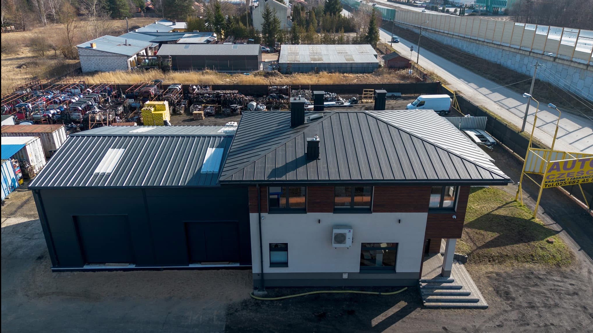 Nowoczesny budynek z ciemną blachą na dachu, widok z góry. Na dachu trzy kominy. Obok budynku skład złomu i części samochodowych. W tle droga i ekrany akustyczne.