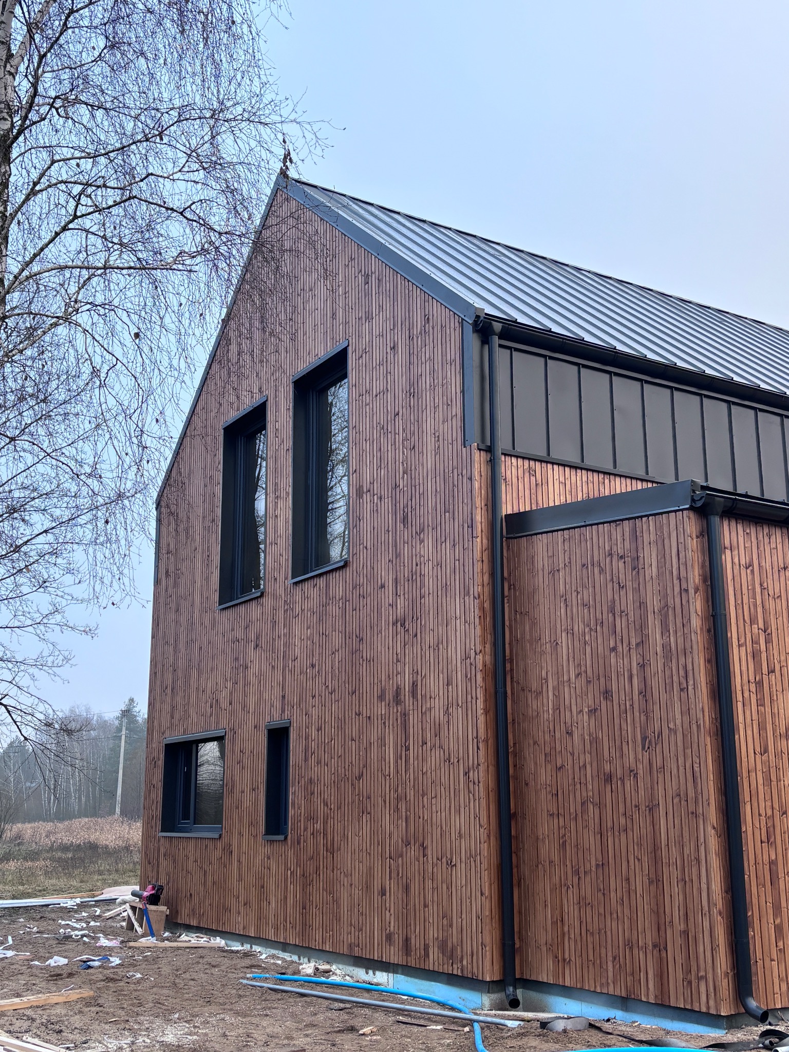 Nowoczesny dom z drewnianą elewacją w pionowe deski i metalowym dachem. Widoczne czarne rynny i okna. Surowy krajobraz w tle.